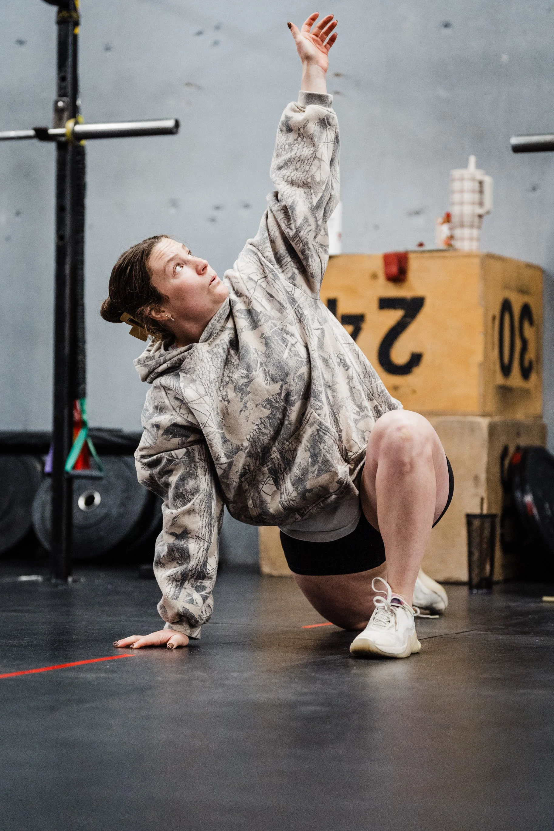 A woman in a camouflage hoodie and shorts performing a stretch balancing on her left hand in a gym with weight equipment and wooden boxes in the background.