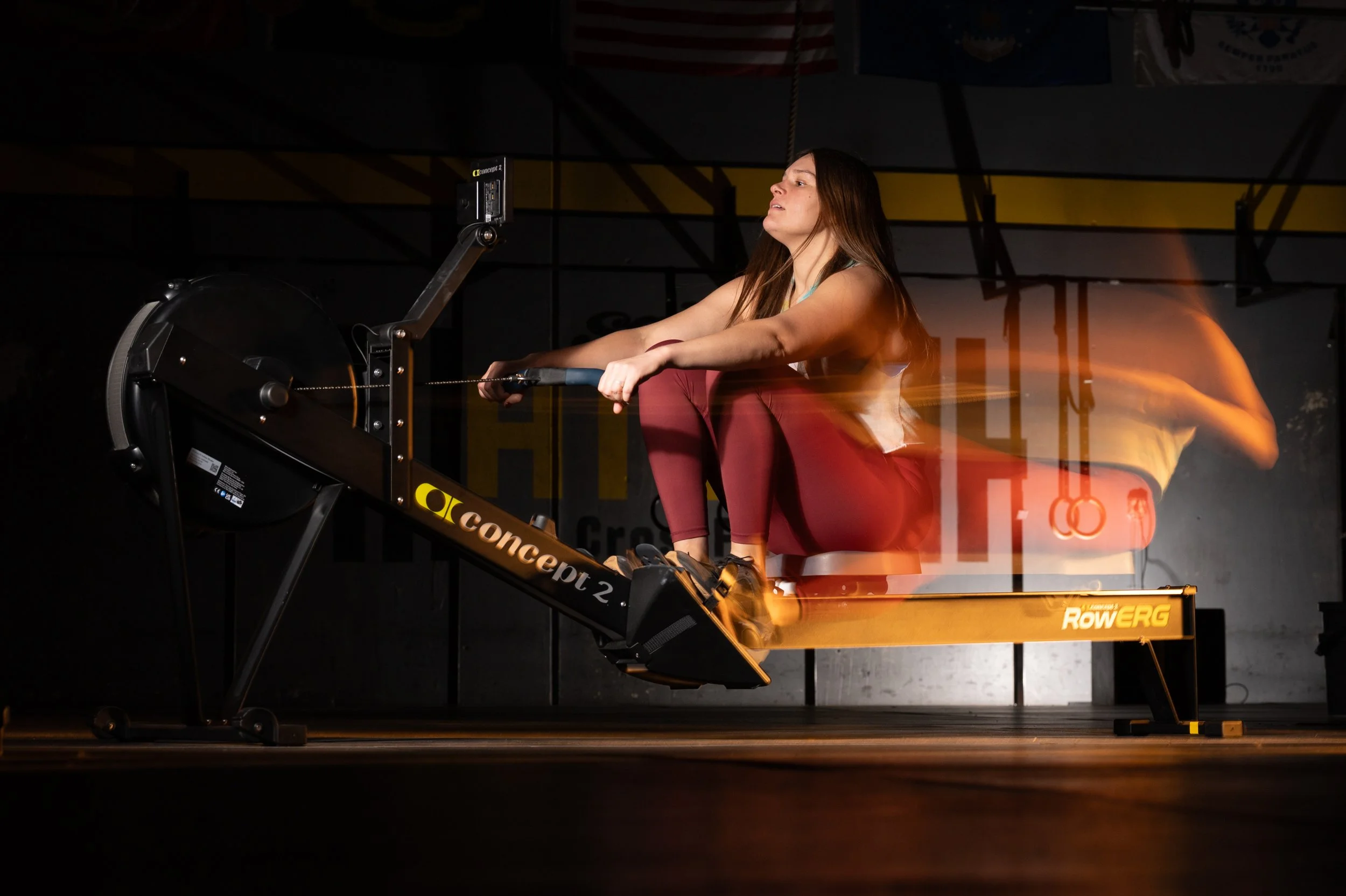A woman using an indoor rowing machine in a gym, with a dark background and gym banners hanging overhead.