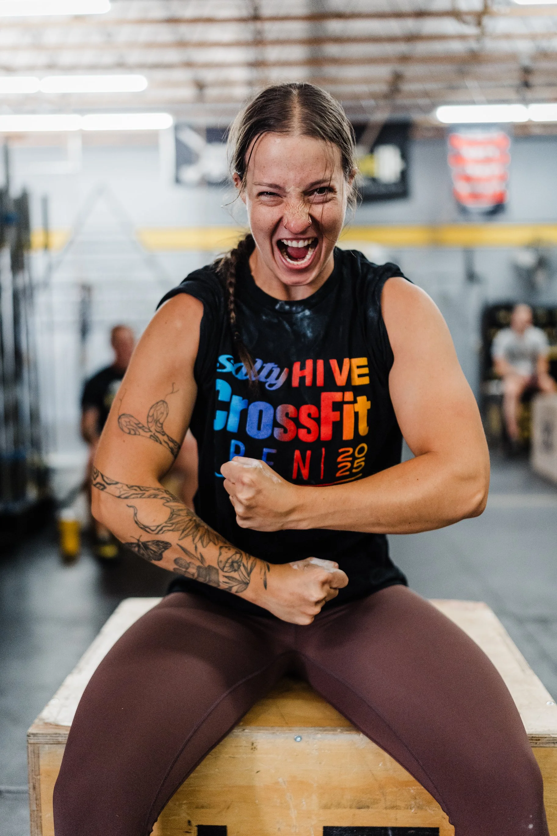 A woman with braided hair and tattoos flexing her arm and smiling in a gym.