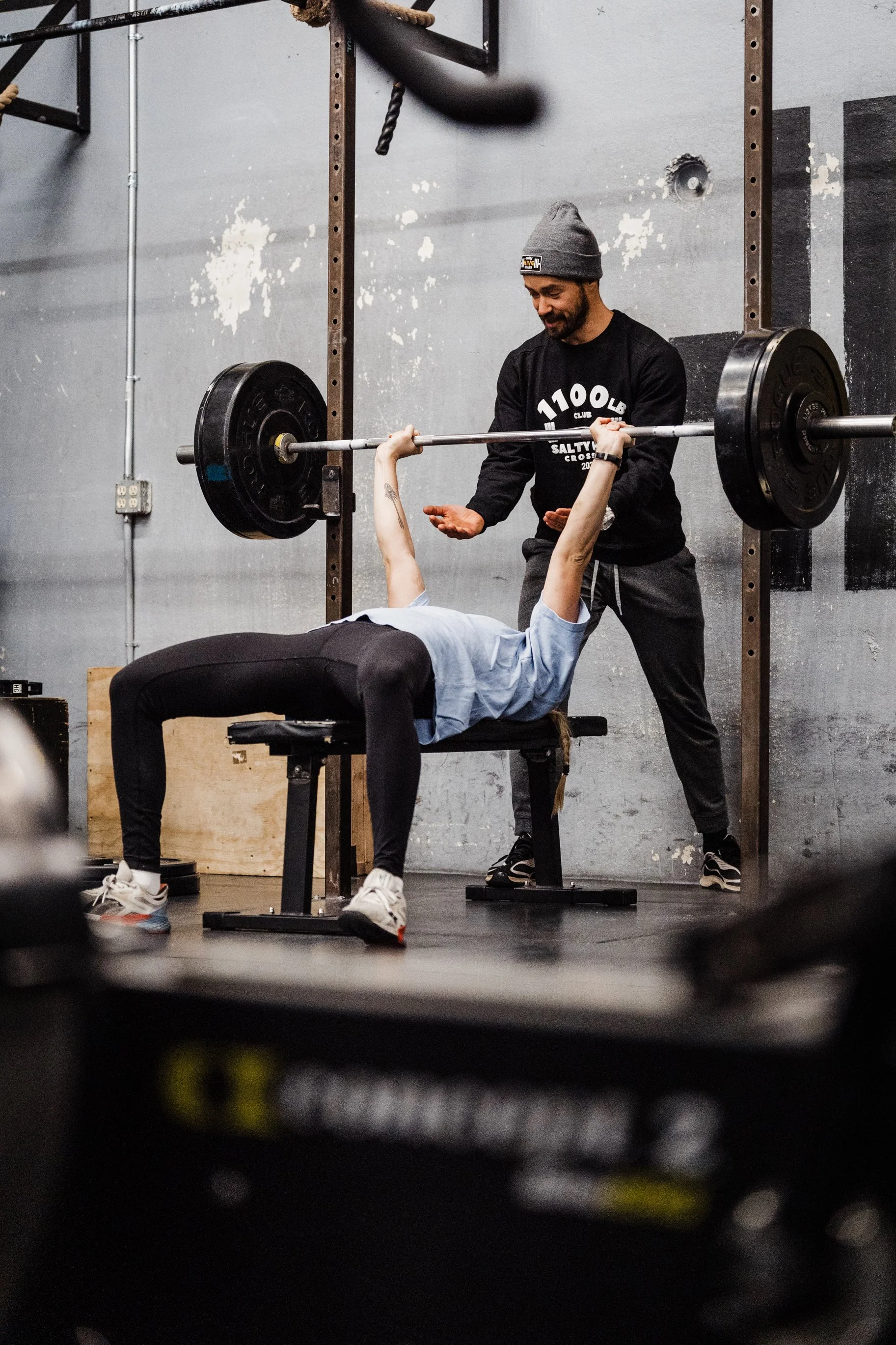 A person lying on a bench lifting a barbell with weights while a trainer supervises in a gym with a gray wall and black markings.