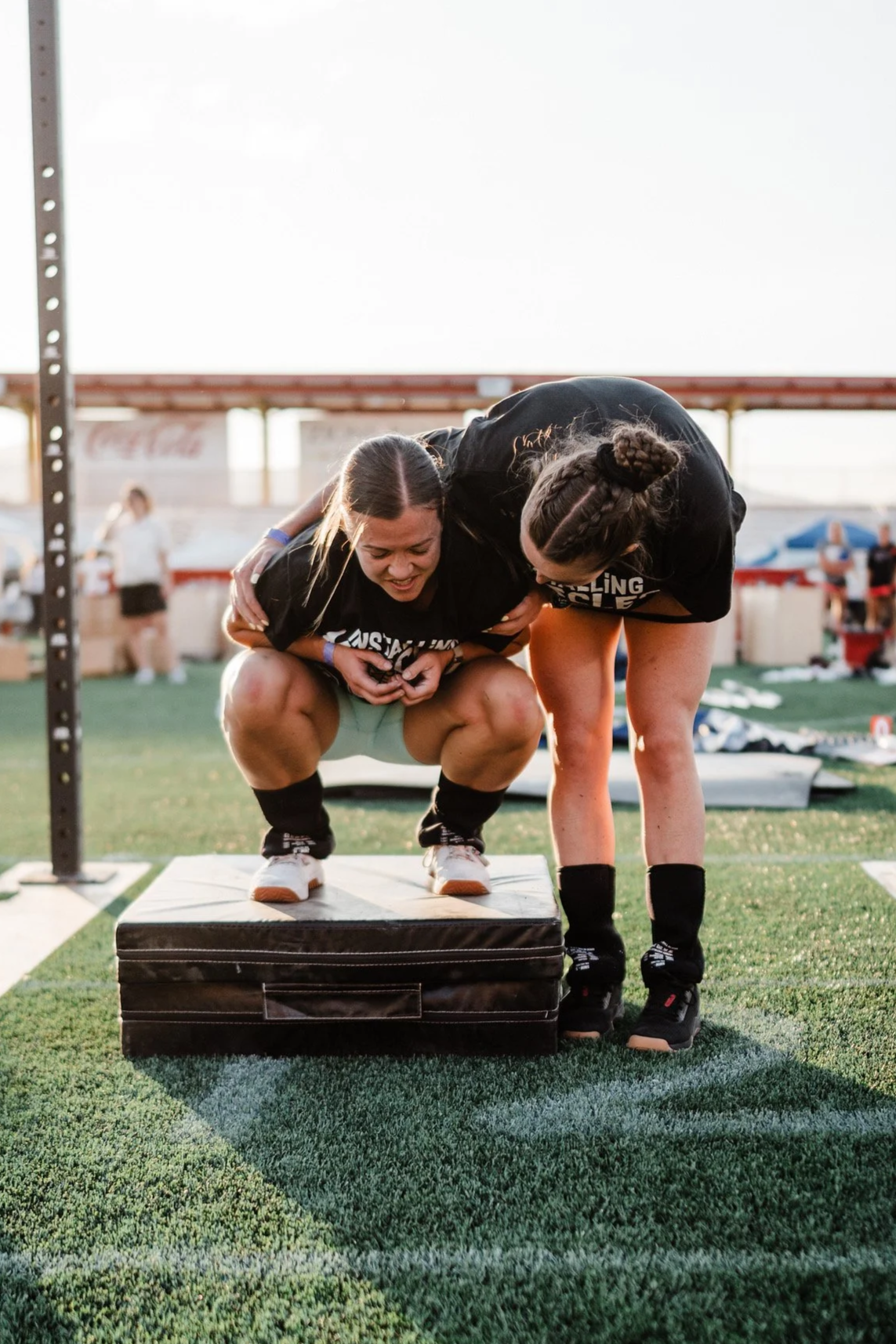 Two female athletes are engaged in a weightlifting event outdoors, with one athlete stabilizing while the other lifts a weight. They are on a sports field with equipment and spectators in the background.