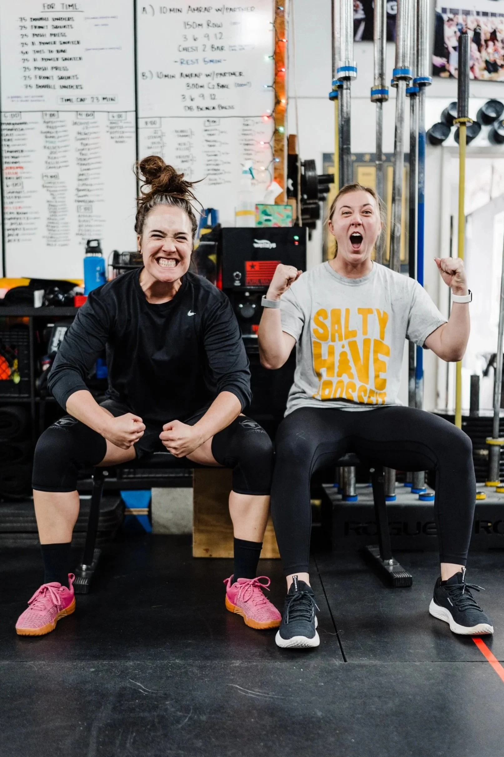 Two women sitting on stools in a gym, flexing their arms and smiling, with a whiteboard and weights in the background.