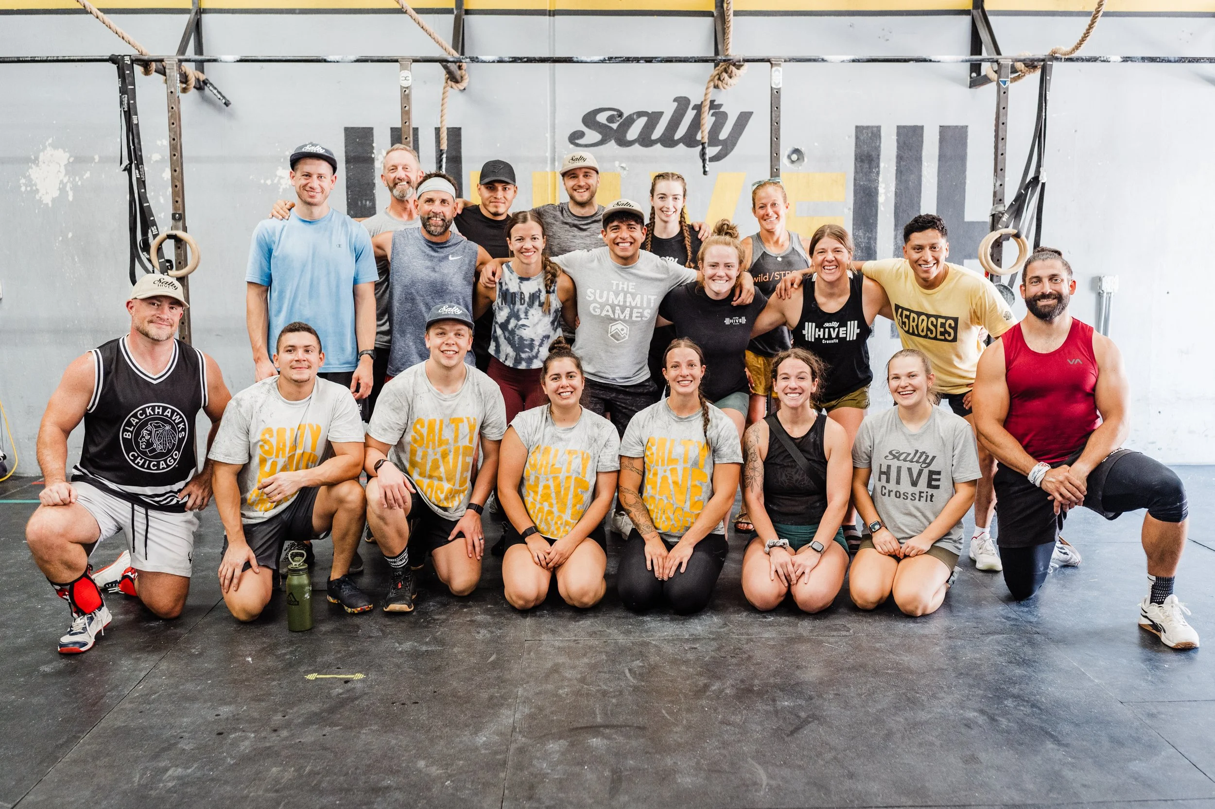 Group of diverse people in athletic clothing posing together in a gym, some sitting and some standing, with gym equipment and a wall with the words 'Salty' and 'LOVE' in the background.