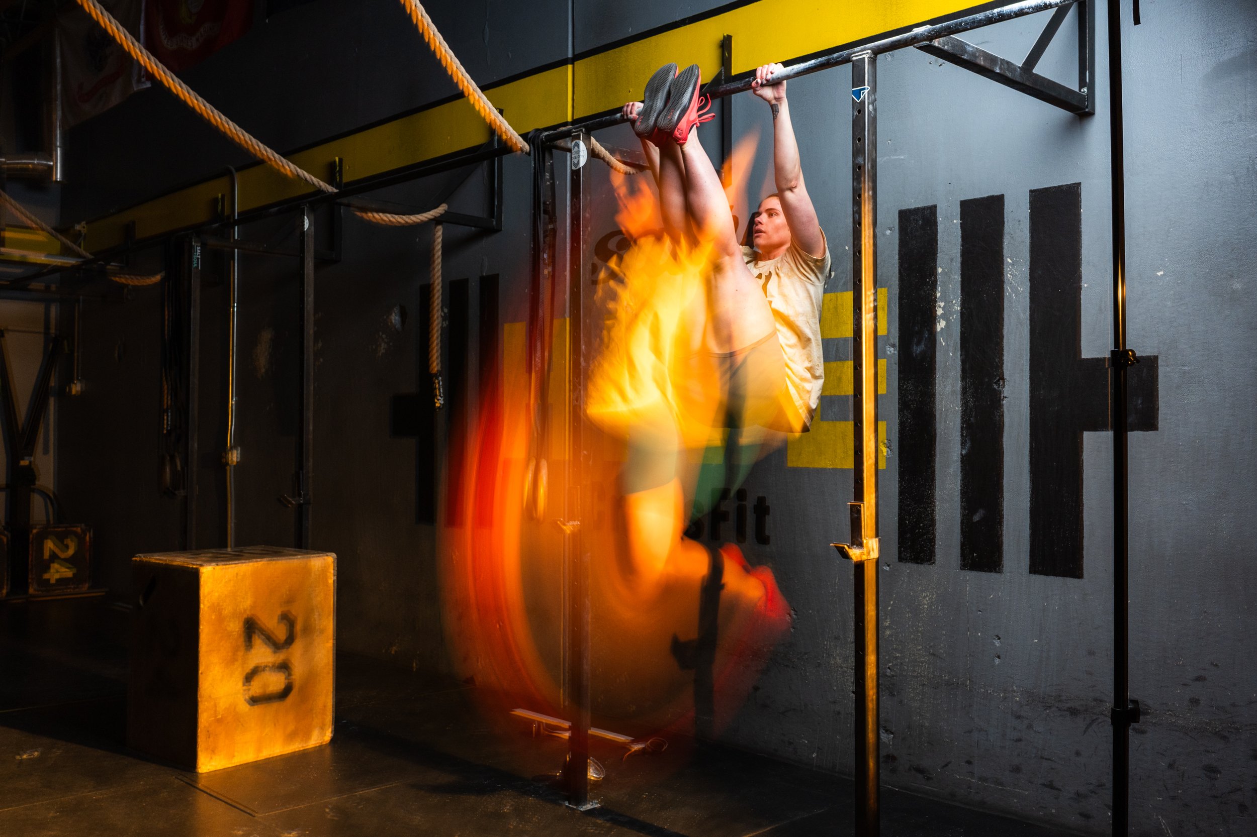 A woman completing toes to bat on a pull-up bar in a gym, with flames near her legs as part of a training exercise or photo effect.