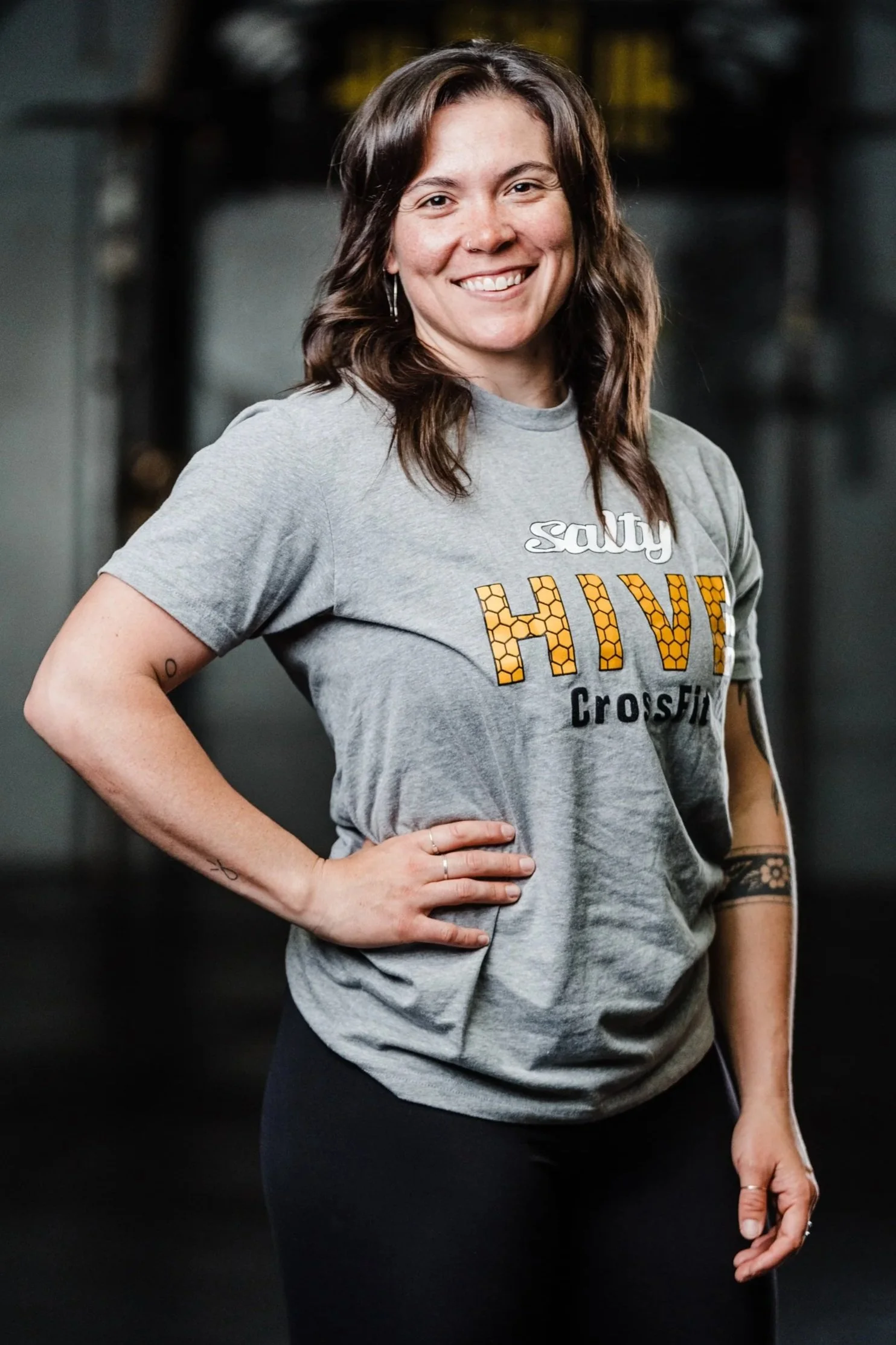 A woman with shoulder-length brown hair smiling, wearing a gray T-shirt with the words 'salty, HIVE, CrossFit' on it, standing with one hand on her hip in a gym setting.
