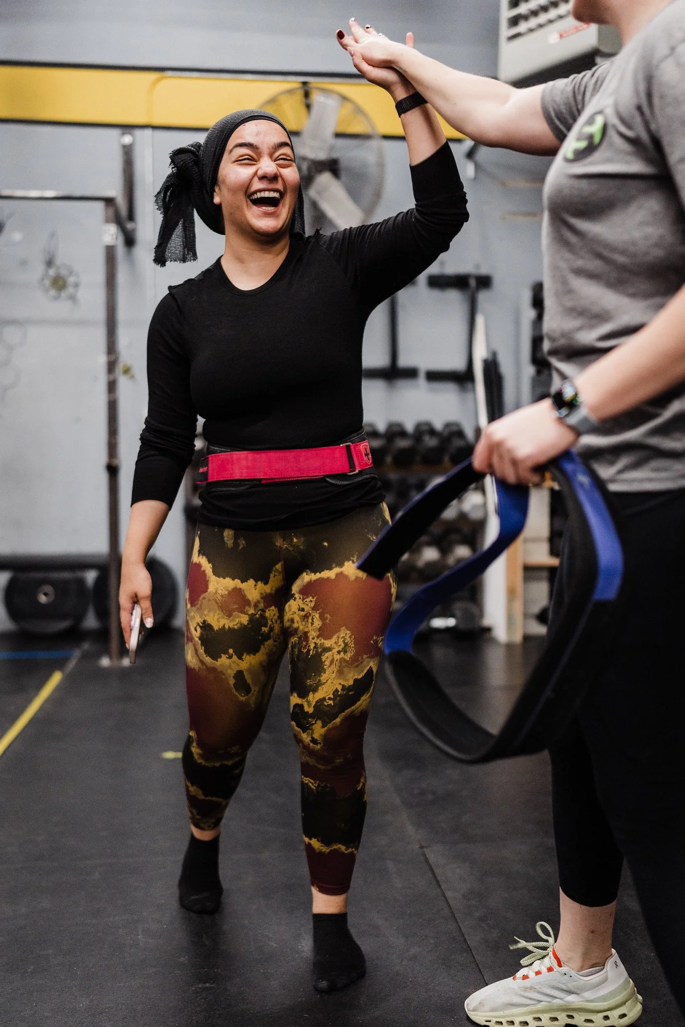 A woman with a headscarf laughing and high-fiving a person at the gym, wearing colorful leggings and black shoes, holding a phone in her hand.