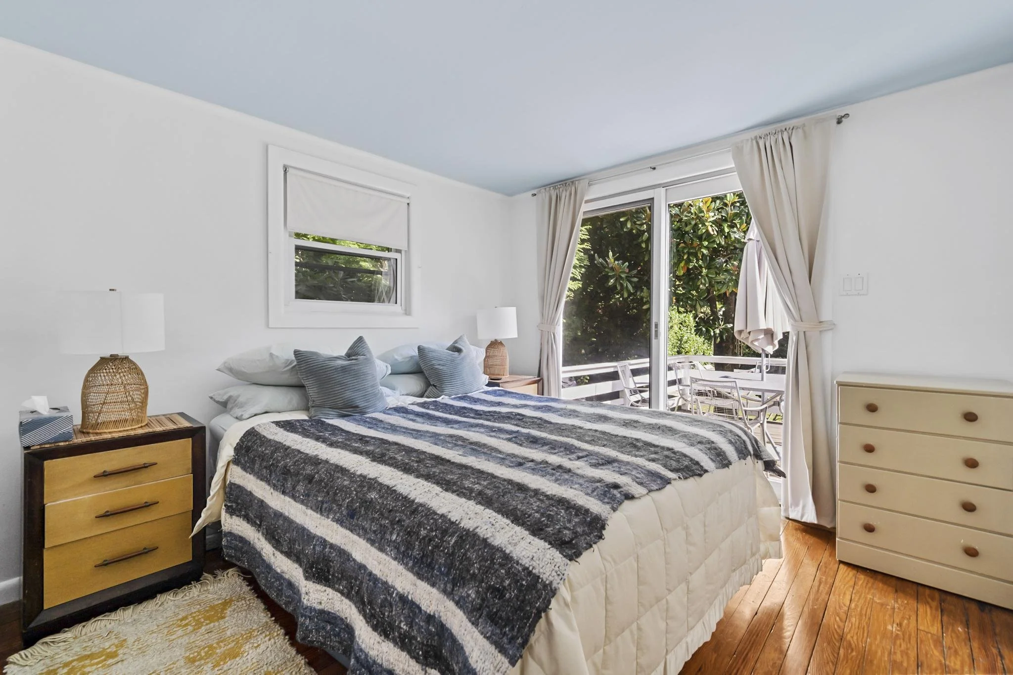 A bedroom with a bed covered in a black, white, and gray striped blanket, two pillows, and two nightstands with lamps on either side. There are large windows with white curtains and a sliding glass door leading to a balcony with outdoor furniture. Th