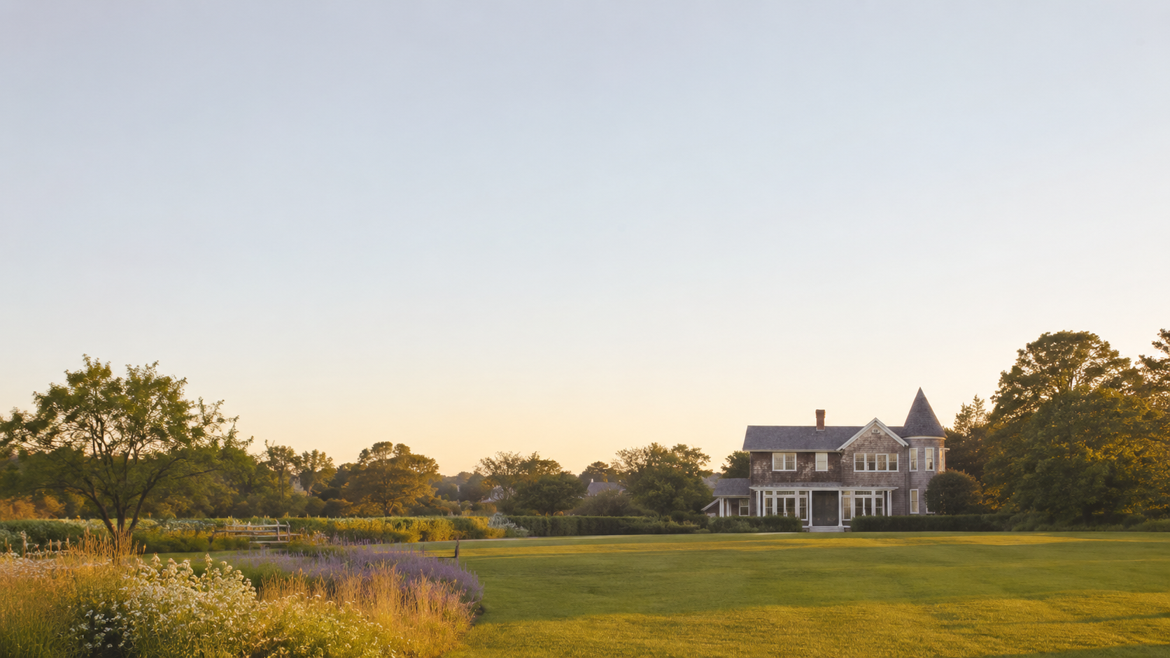 A large, historic house with a turret, surrounded by a well-maintained lawn and garden, under a clear sky at sunset.