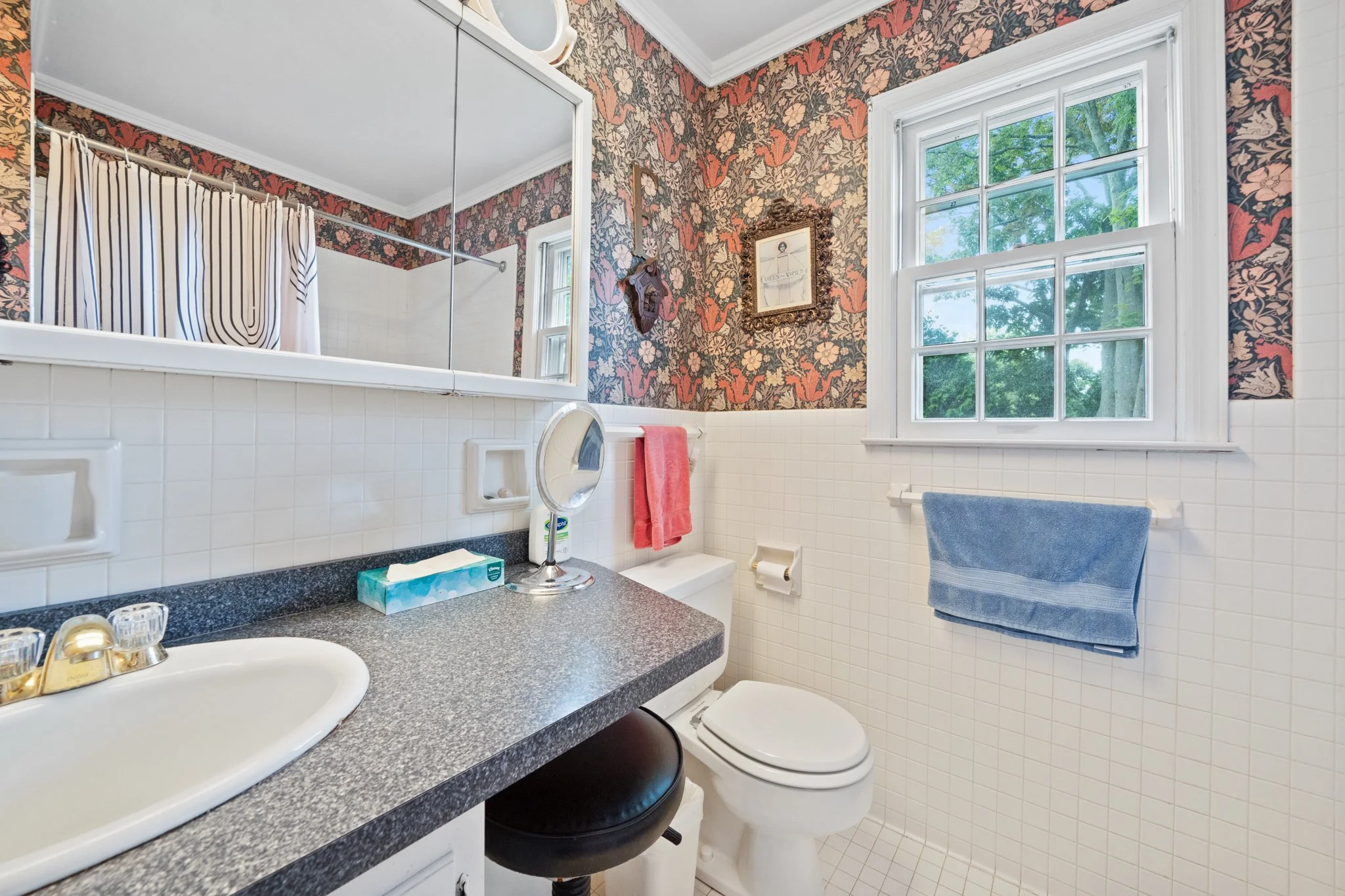 Small bathroom with floral wallpaper, a window showing green trees outside, a white tiled wall, a toilet with a blue towel, a countertop with tissues, a mirror, a small sink, and a washing machine.