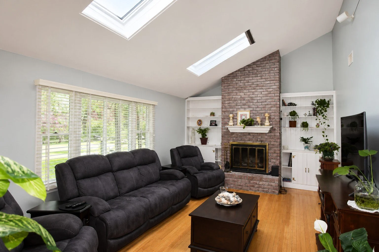 Living room with black recliner sofas, brick fireplace with candles, white built-in shelves with plants and books, and a window with white blinds. Skylights on the ceiling and hardwood floors.
