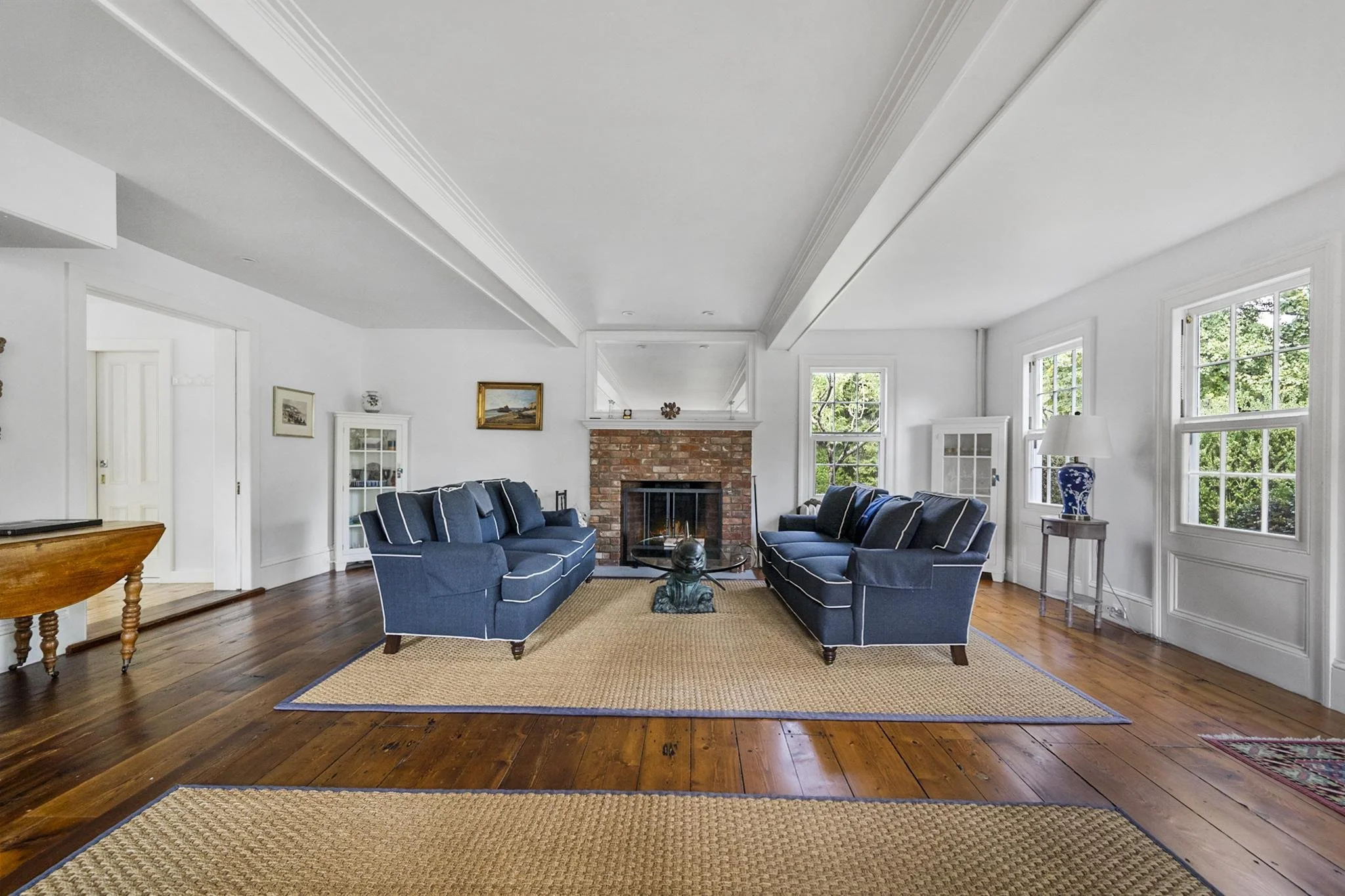 Living room with white walls, hardwood floors, blue sofas, a brick fireplace, and multiple windows letting in natural light.