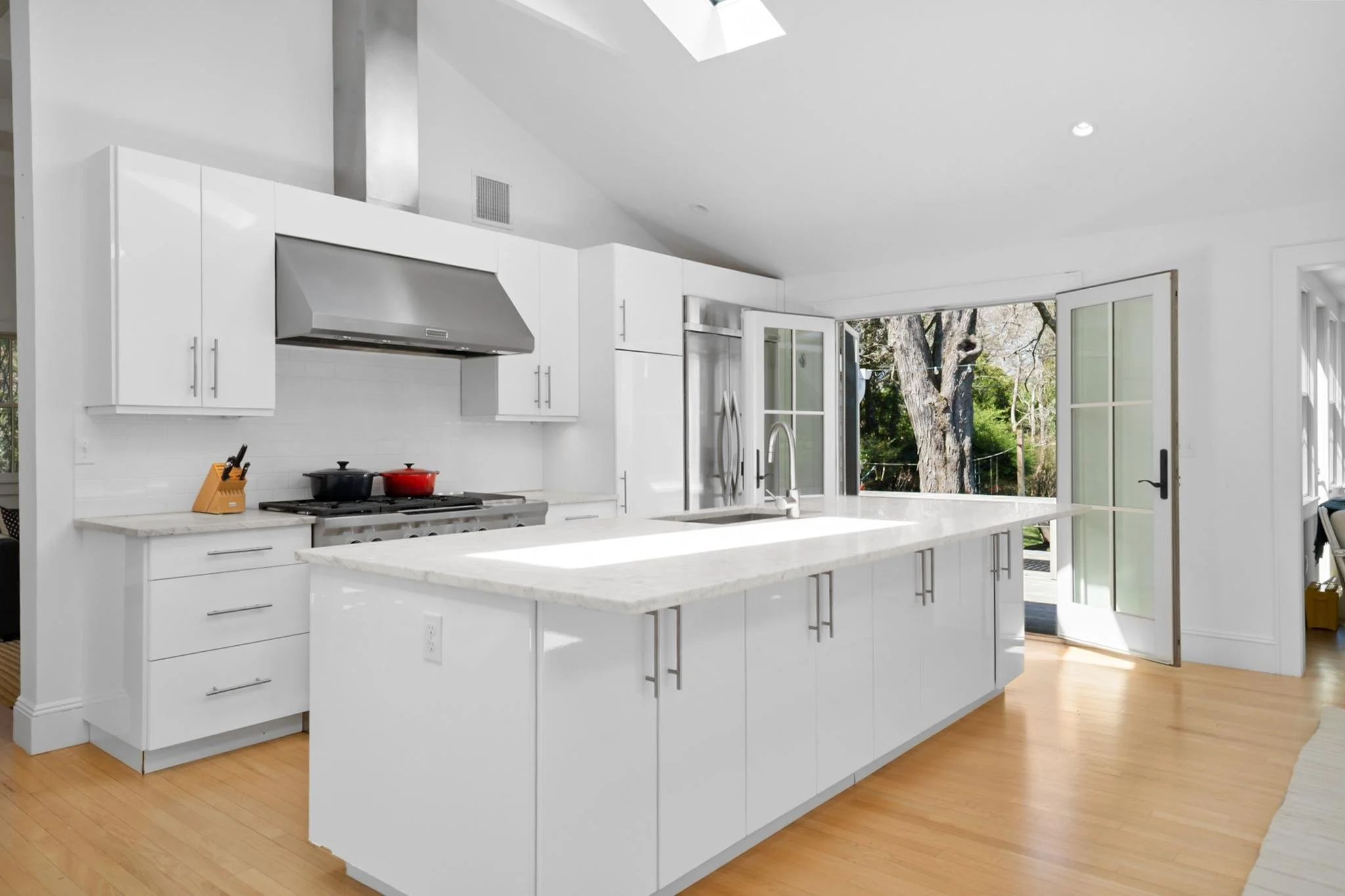 Modern white kitchen with large island, stainless steel appliances, and open double doors leading to outdoor trees.