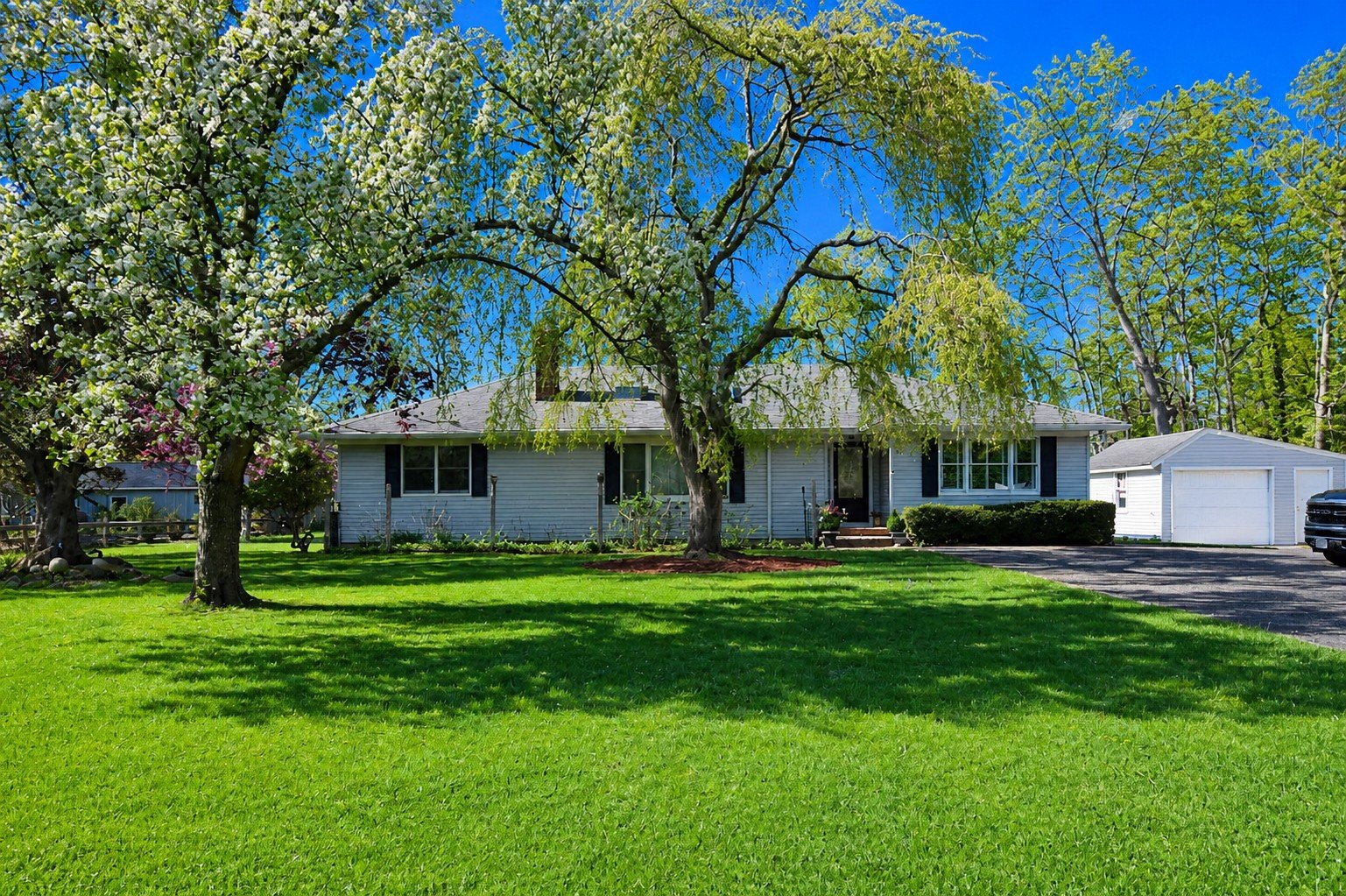 Front view of a house with manicured lawn, large mature trees with blooming flowers, and a detached garage on a bright sunny day with a clear blue sky.