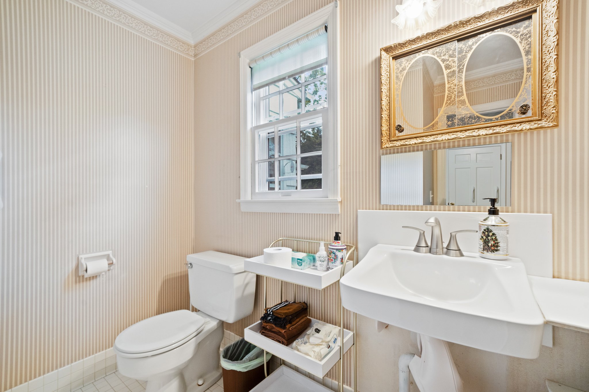A small bathroom with a window, a toilet, a white shelf with toiletries, and a white porcelain sink with a silver faucet, decorated with beige and white striped wallpaper and a gold-framed mirror.