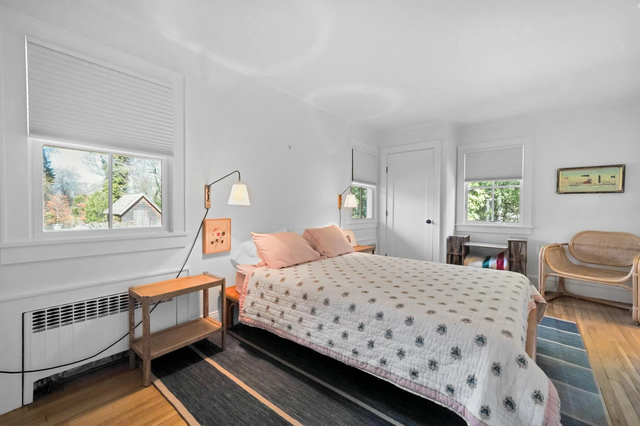 A bedroom with white walls and windows. There is a bed with pink pillows and a patterned quilt. Two wall-mounted lamps are beside the bed. A wooden side table is next to the bed. There is a framed picture on the wall, and a wicker chair and some stor