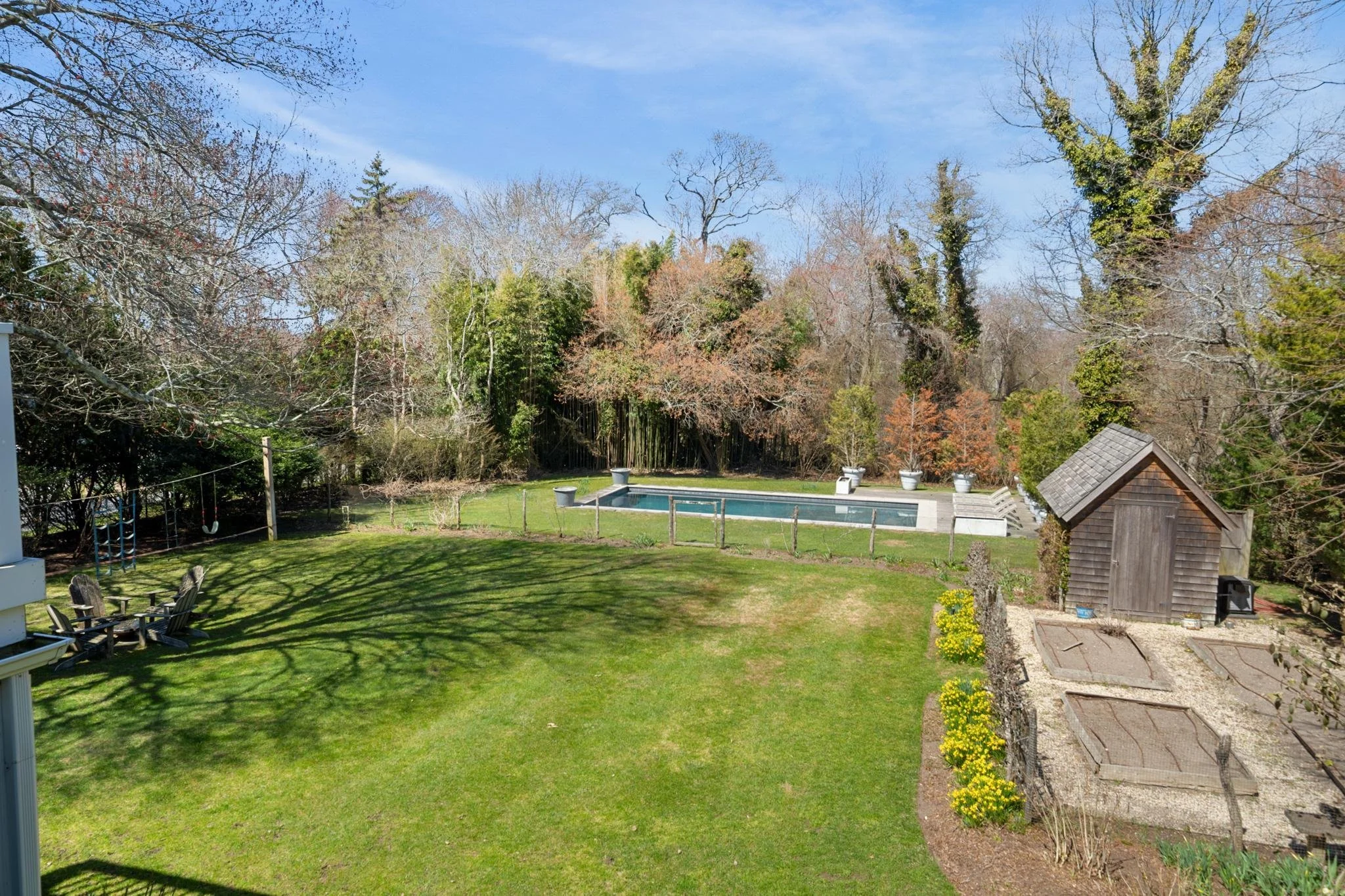 Backyard with green grass, wooden patio furniture, children's swing set, swimming pool, small wooden shed, and trees with some leaves and some bare branches, under a clear blue sky.