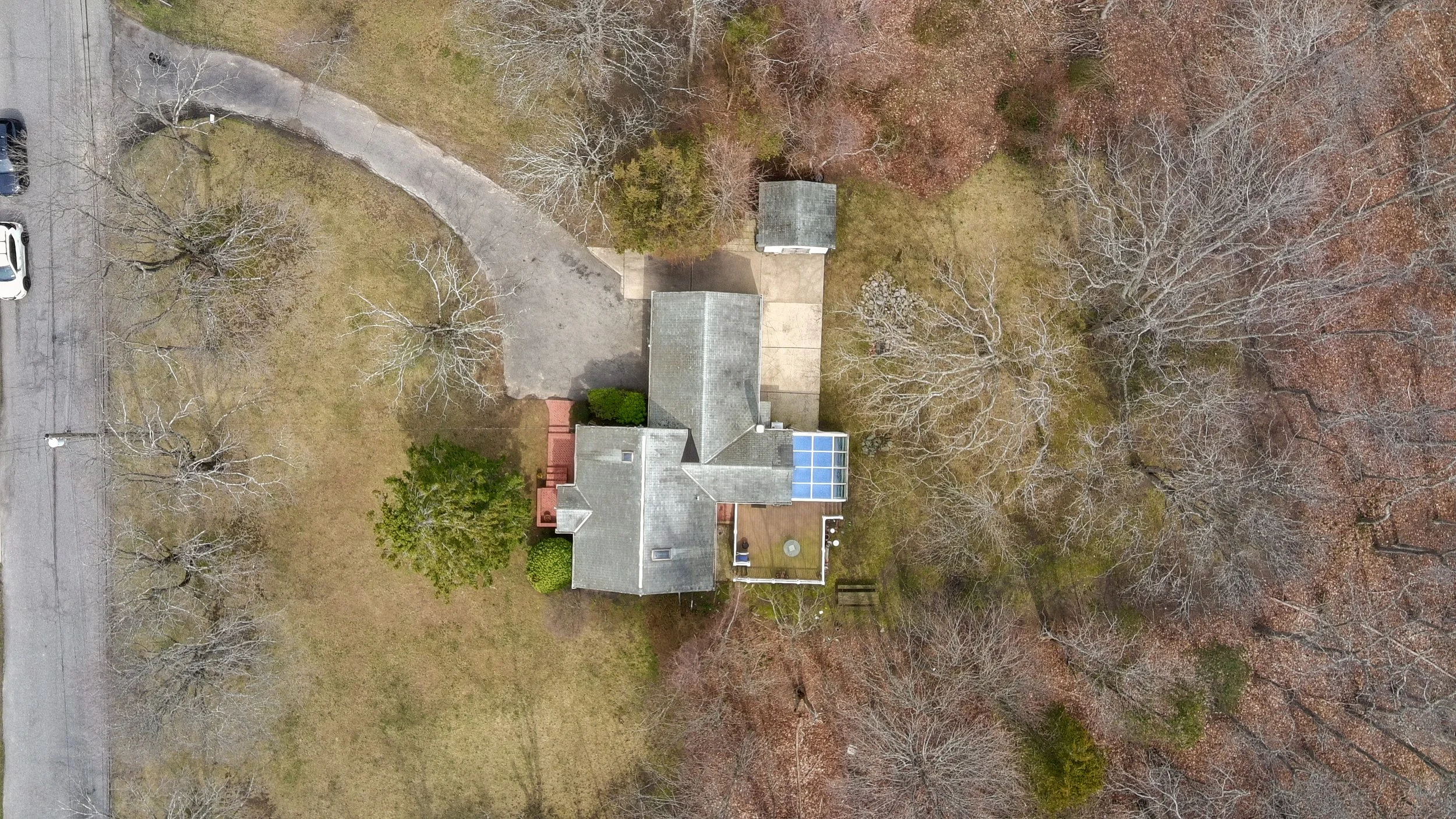 Aerial view of a house with a gray roof, small porch, and a sunroom with solar panels, surrounded by leafless trees and a grassy yard in winter. A curved driveway runs from the street to the house, with a parking area at the end.