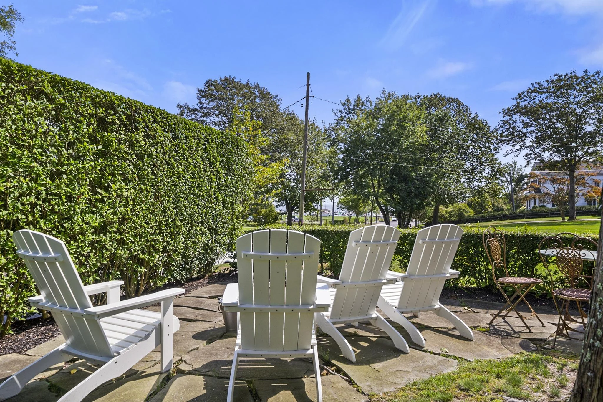 White wooden Adirondack chairs arranged in a semi-circle on a patio, overlooking a lush garden with a hedge, trees, and a clear blue sky.