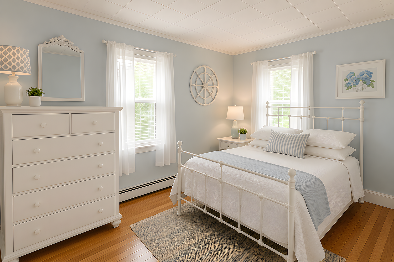 A bright bedroom with light blue walls, white furniture, and hardwood floors. The bed has white bedding and a striped pillow, flanked by two windows with sheer curtains. There is a white dresser, a mirror, a bedside table with a lamp, and decorative 