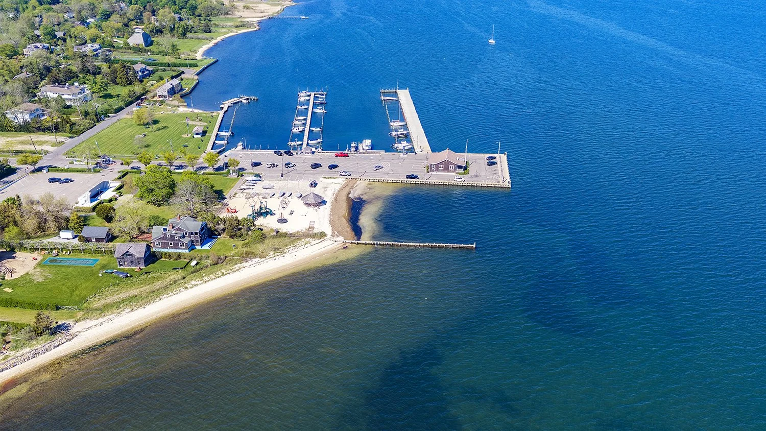 Aerial view of a coastal marina with boats docked, a parking lot, and residential houses along the shoreline and a sandy beach.
