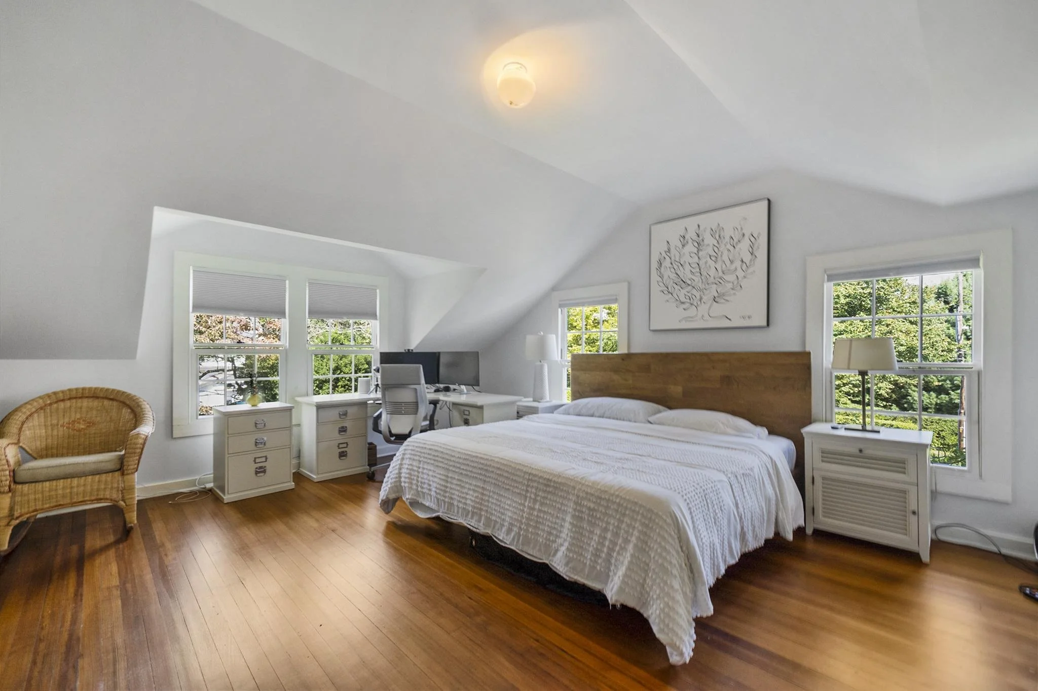 Bright bedroom with white walls, a white bed with a wooden headboard, multiple windows with blinds showing greenery outside, a wicker chair, a white desk with a computer, and framed artwork of a tree on the wall.
