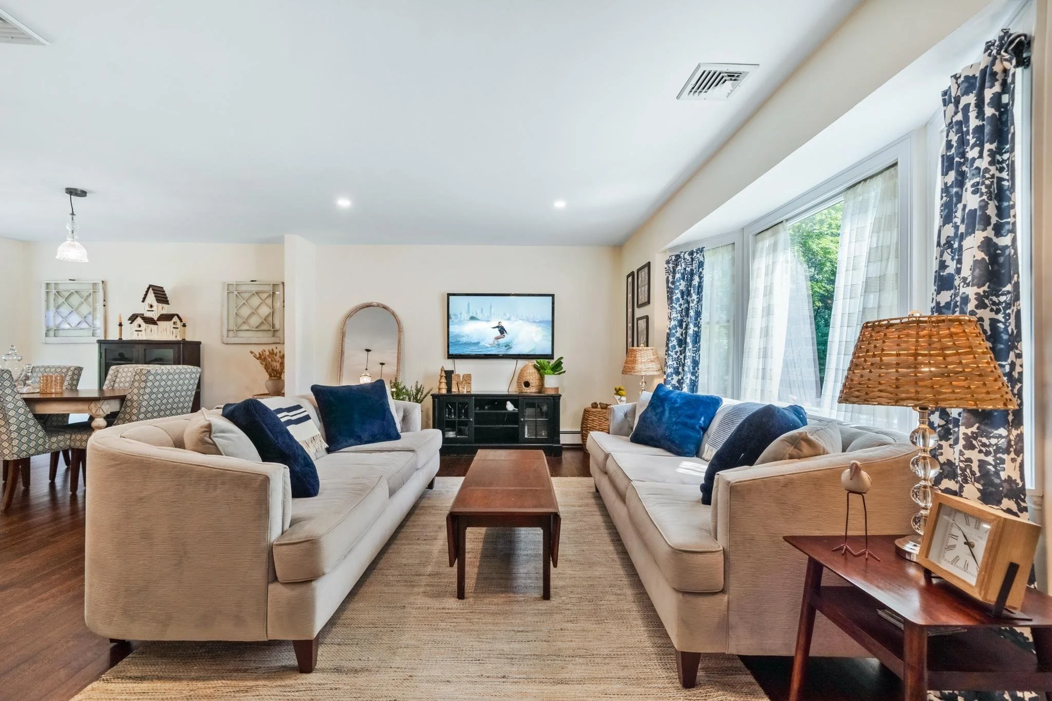 Living room with two beige sofas with blue cushions, a wooden coffee table, a black TV stand with a TV, large windows with blue and white floral curtains, a wicker floor lamp, framed pictures on the wall, and a dining area in the background.