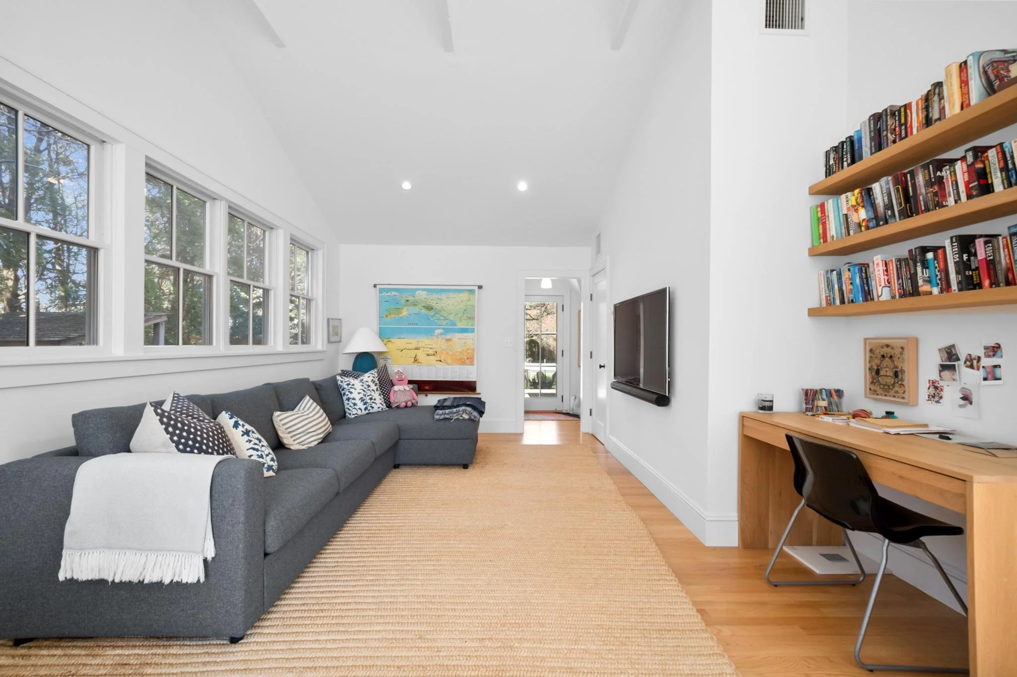 A living room with a gray sofa, patterned pillows, a side table, a lamp, a colorful beach-themed wall map, large windows, and a flat-screen TV on the wall. There's a wooden desk with a black chair and a shelf filled with books on the right.