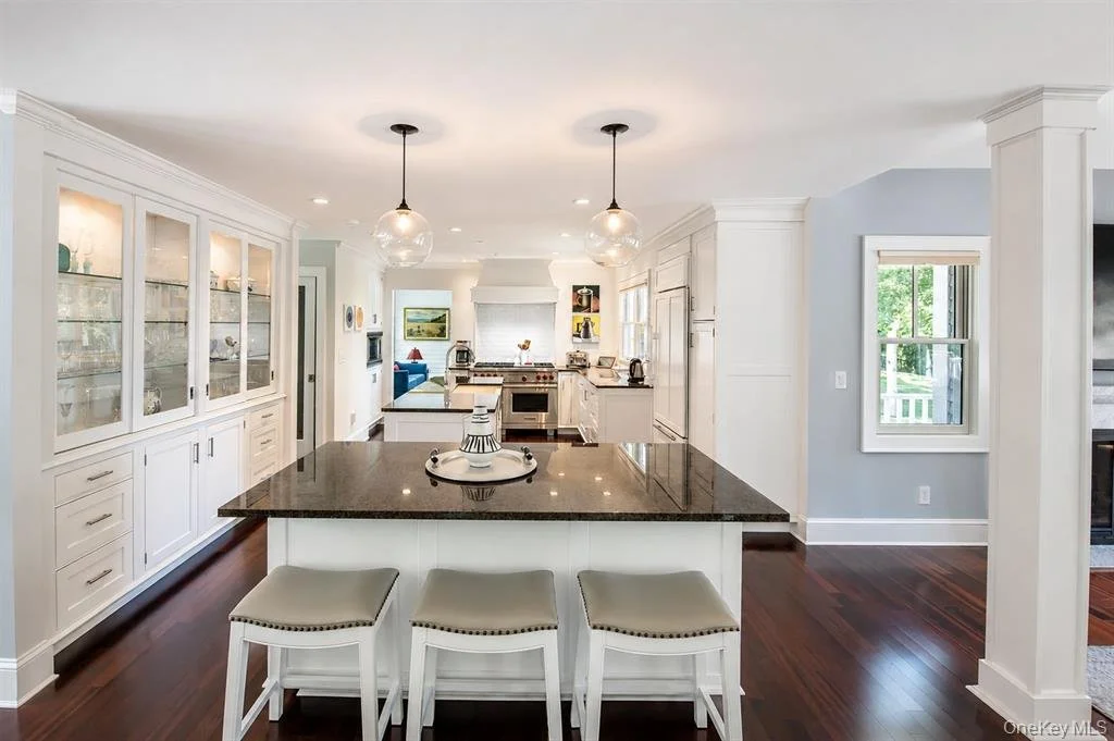 Modern kitchen with white cabinets, dark granite island, three beige barstools, pendant lights, and hardwood floors.