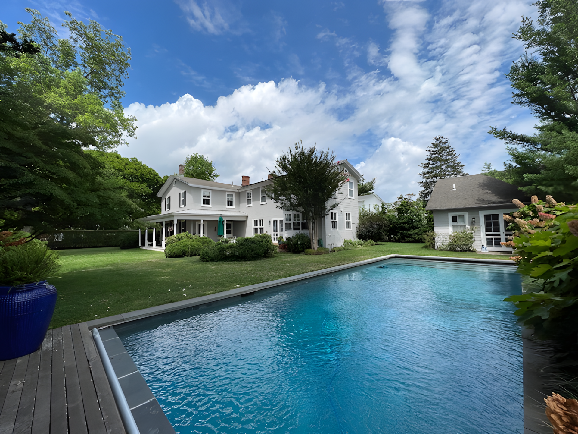 A backyard with a swimming pool, surrounded by green grass, trees, and bushes. There are two white houses with multiple windows and a porch. The sky is blue with scattered clouds.