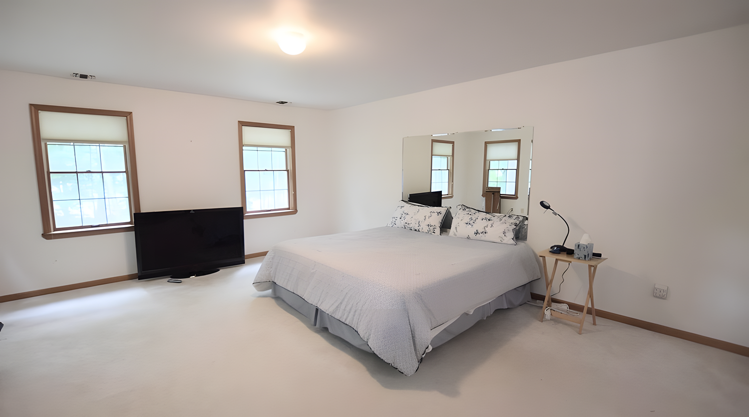 Bedroom with white walls, three windows, a bed with white and black floral pillows, a side table with a lamp, a TV on the floor, and a mirror on the wall.