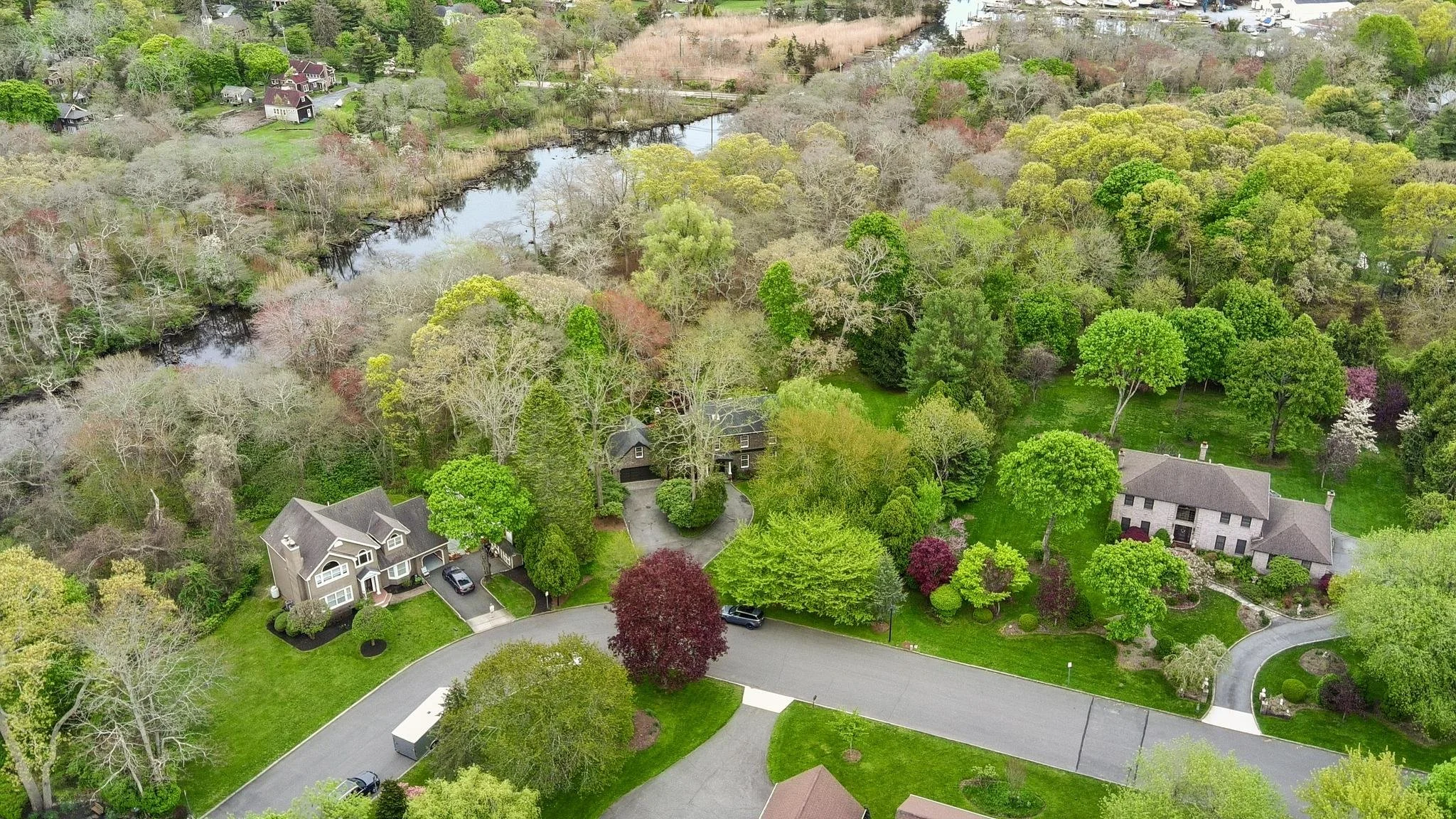 Aerial view of a residential neighborhood with houses, lush green trees, and a small winding road surrounded by greenery and a small river or creek.