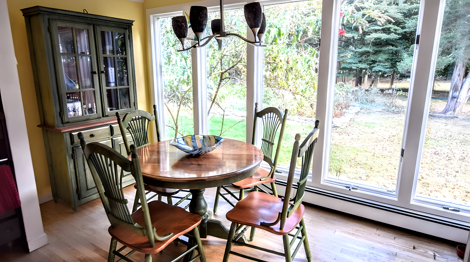 Interior of a bright dining room with a round wooden table, six chairs, a blue glass bowl, a large window showing an outdoor yard with trees and grass, and a green cabinet against a yellow wall.