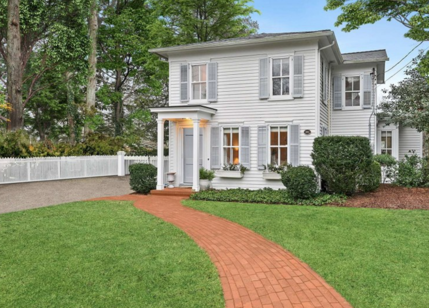 White two-story house with gray shutters, front porch with columns, and a curved brick walkway surrounded by green lawn and bushes.