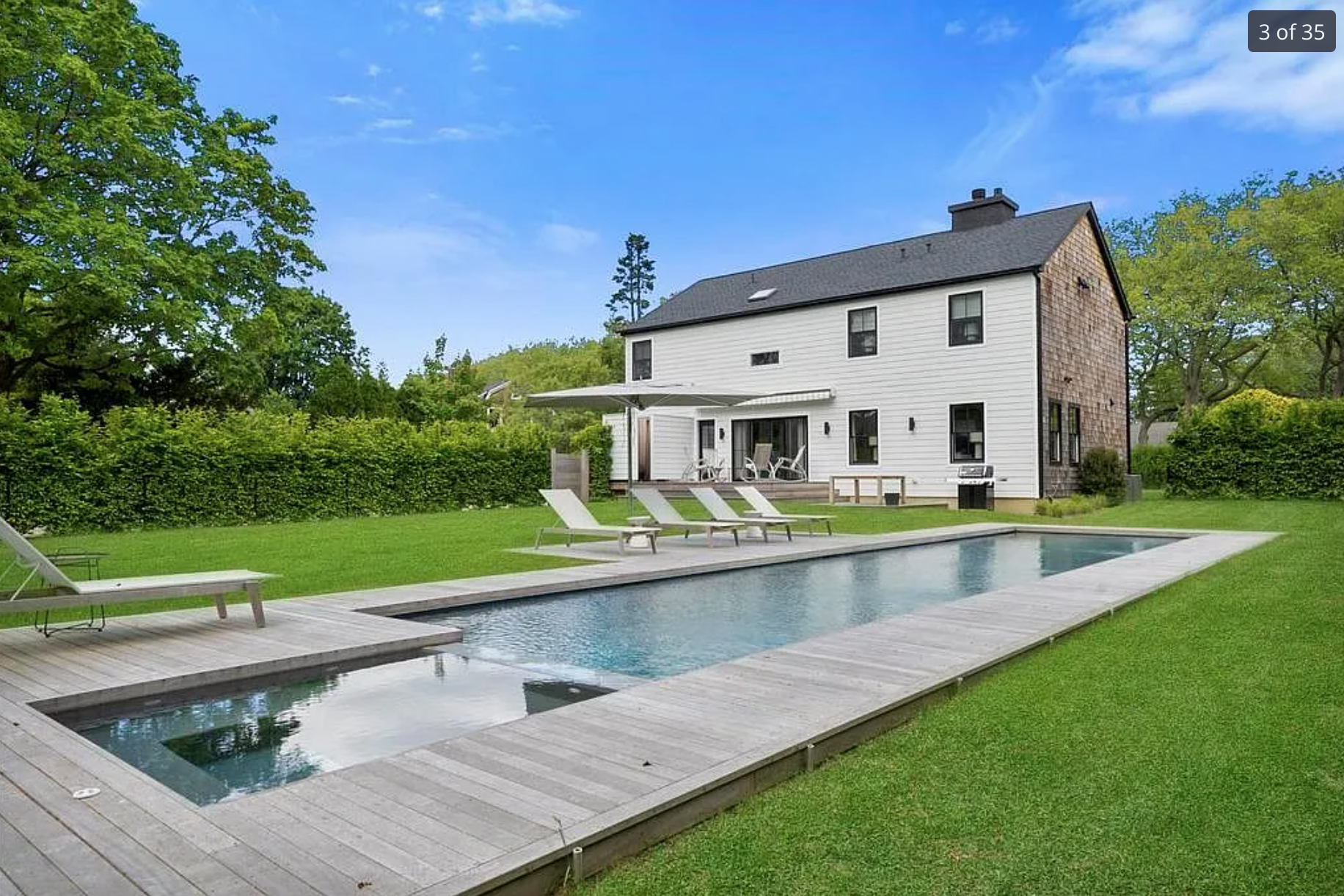 A backyard with a rectangular swimming pool, a wooden deck, and several lounge chairs. There is a modern two-story house with white siding, black window frames, and a covered patio area with outdoor furniture. Green trees and bushes surround the yard, and the sky is mostly clear with some clouds.