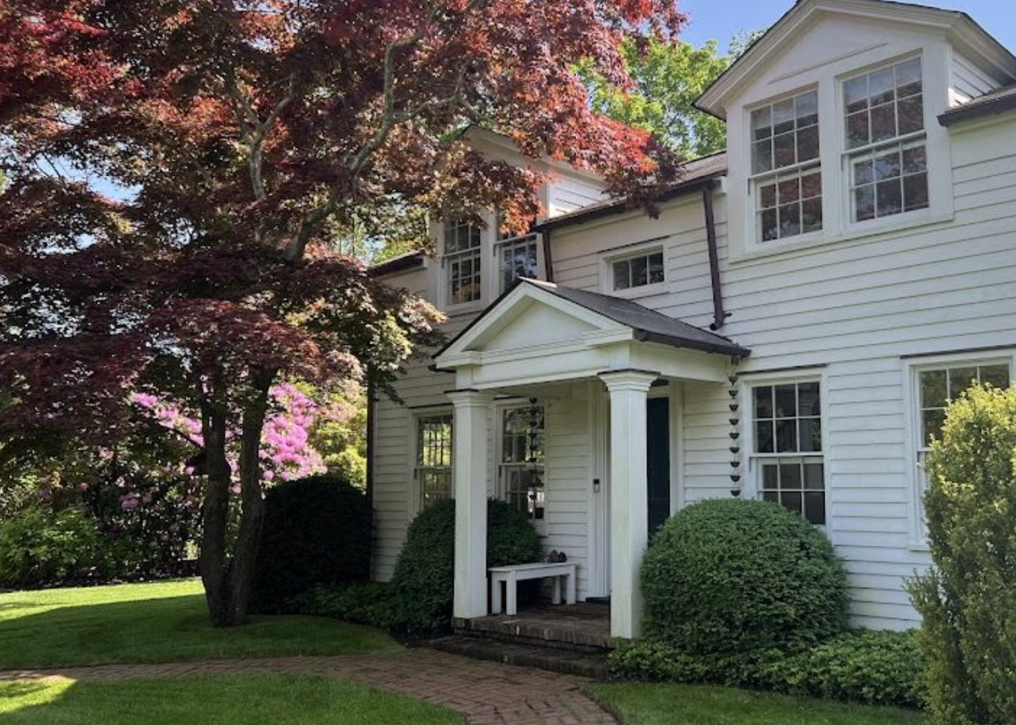White two-story house with a gabled porch, surrounded by well-maintained garden with green bushes and trees with pink and red leaves.
