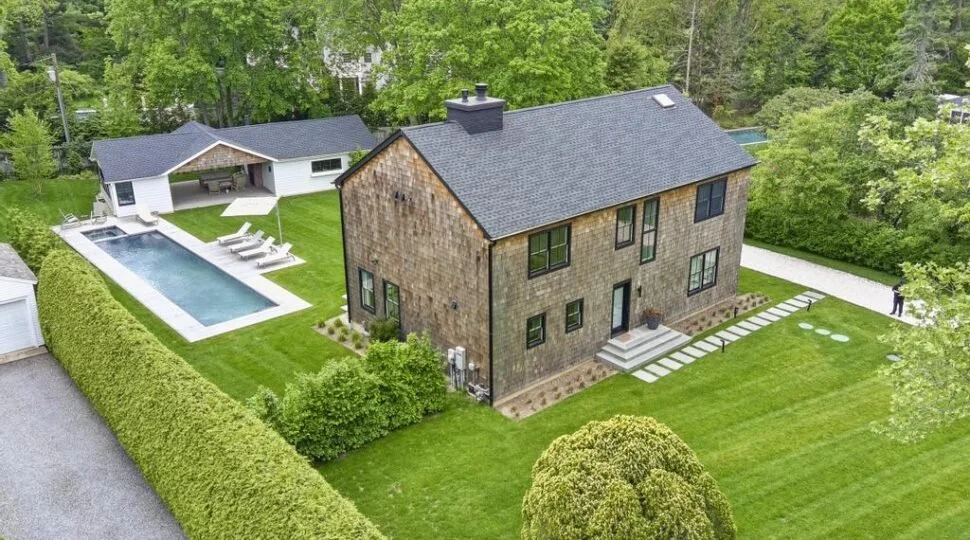 A backyard scene featuring a modern house with a dark roof, a swimming pool with lounge chairs, a detached covered patio, and lush green trees surrounding the area.