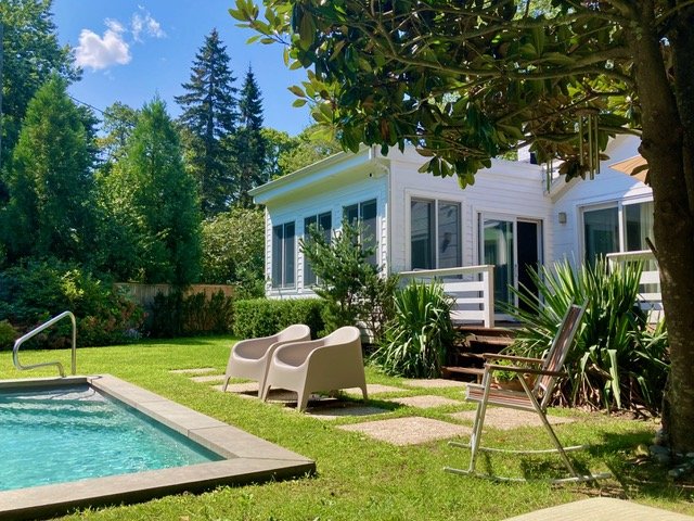 A backyard with a swimming pool, patio furniture, and a white house with large windows, surrounded by lush green trees and plants.