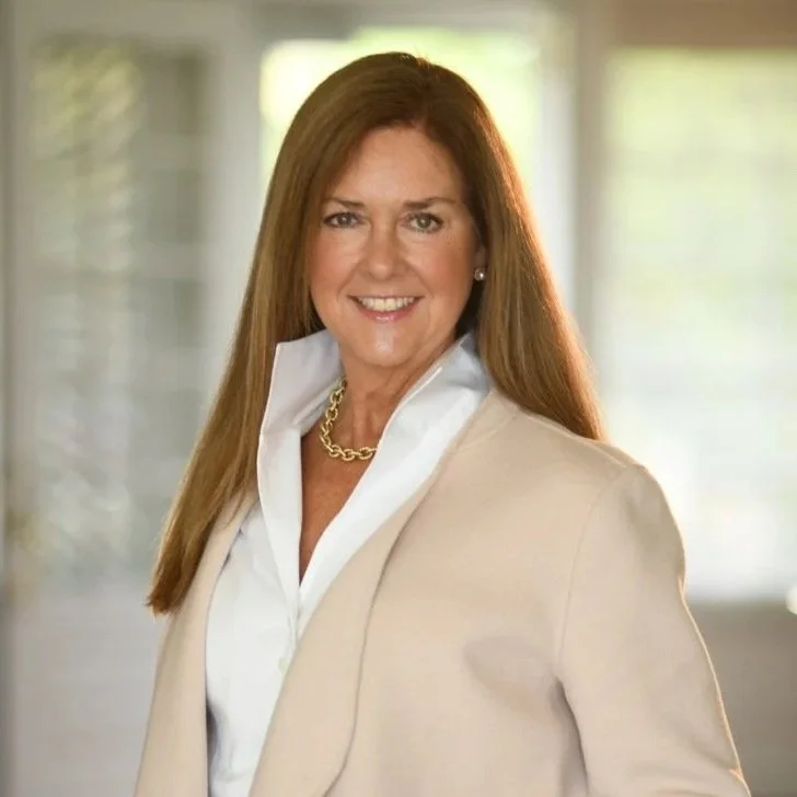 A woman with long, reddish-brown hair smiling, wearing a white shirt, beige blazer, and a gold chain necklace, standing in an indoor setting with blurred windows in the background.