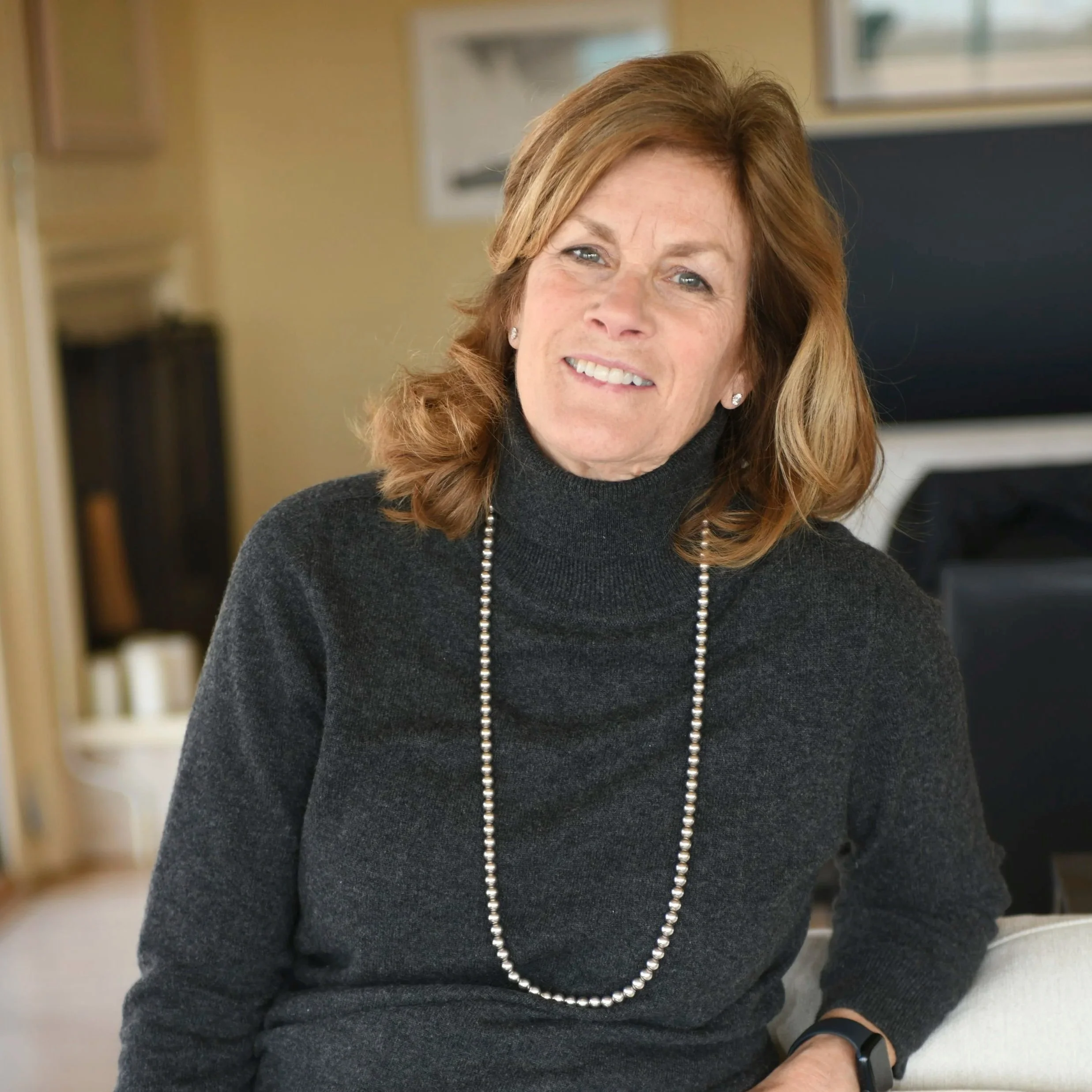 Close-up of a smiling middle-aged woman with auburn hair, wearing a black turtleneck sweater, a pearl necklace, and pearl earrings, indoors with a blurred background.