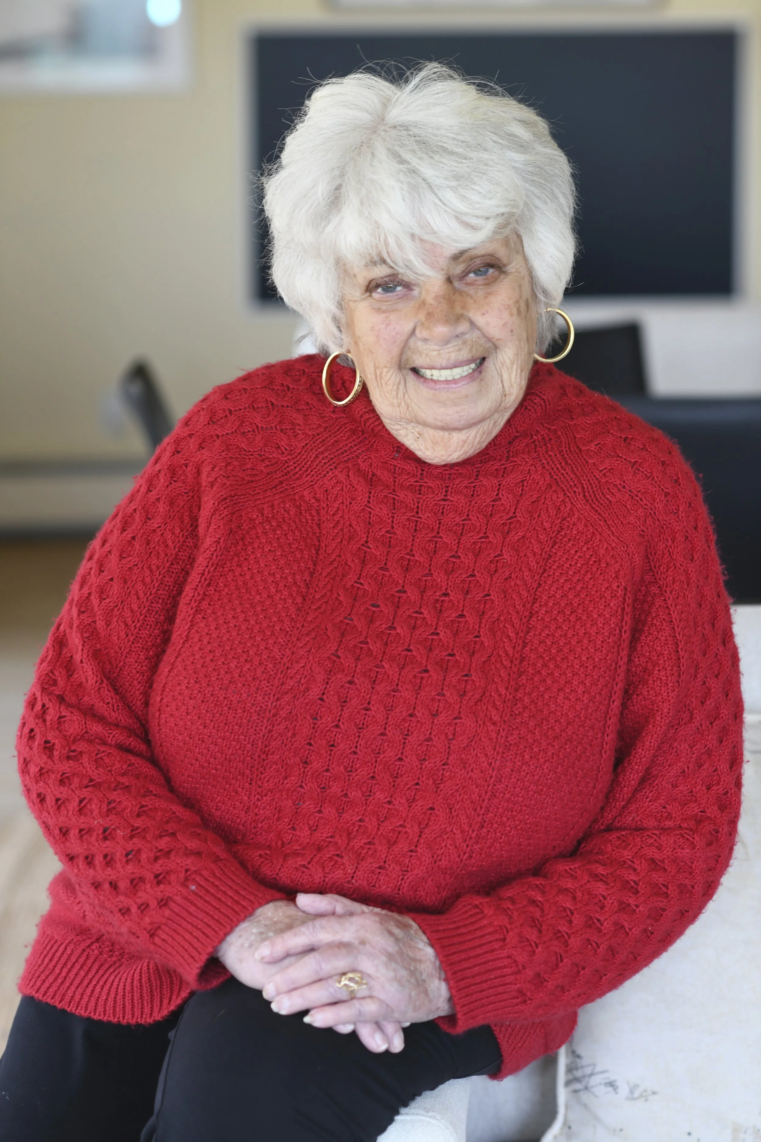 An elderly woman with white hair wearing a red knit sweater and gold hoop earrings, smiling and sitting in a room with a black computer monitor in the background.