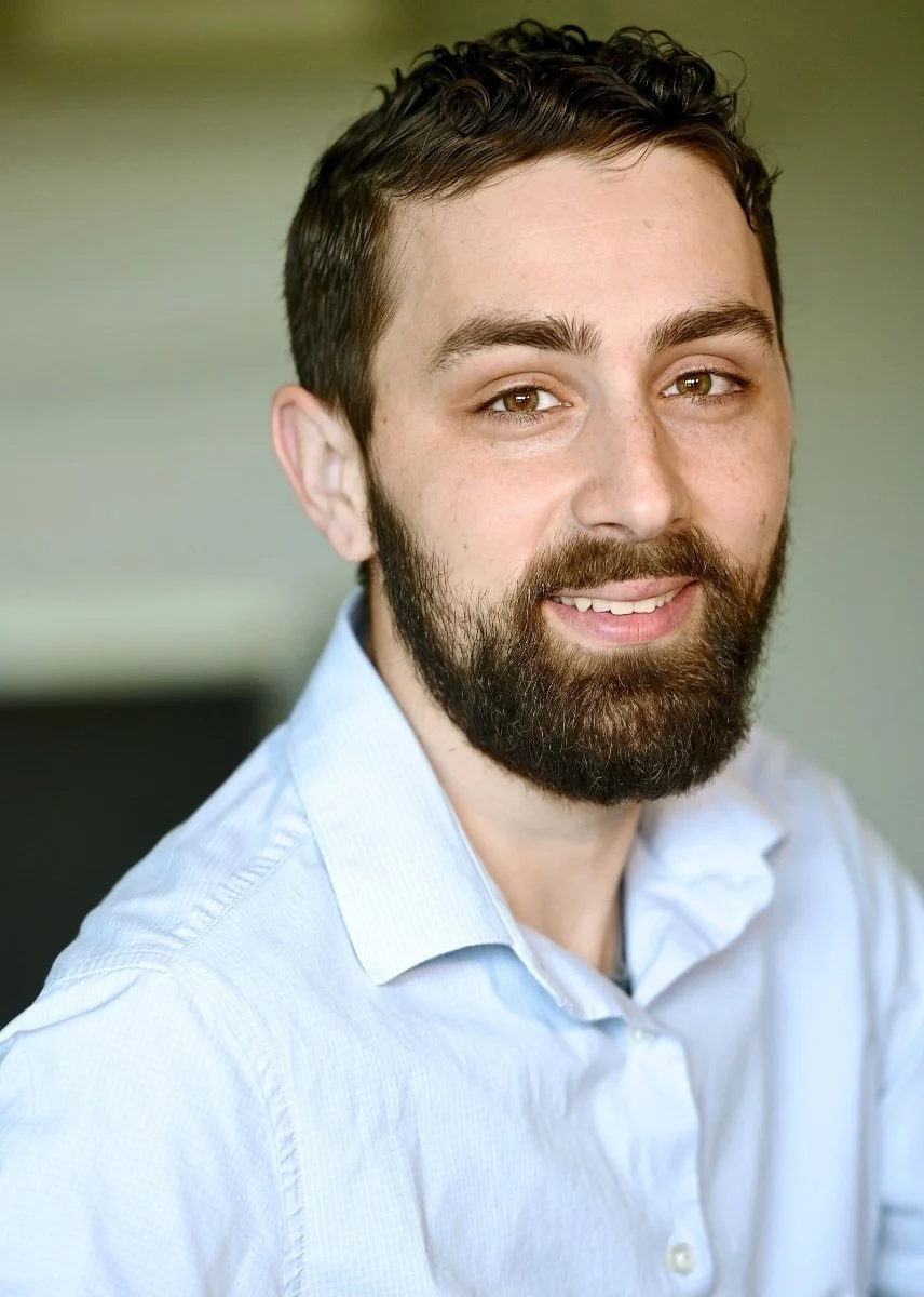 Kyle with dark hair and a beard, wearing a light blue shirt, smiling at the camera.