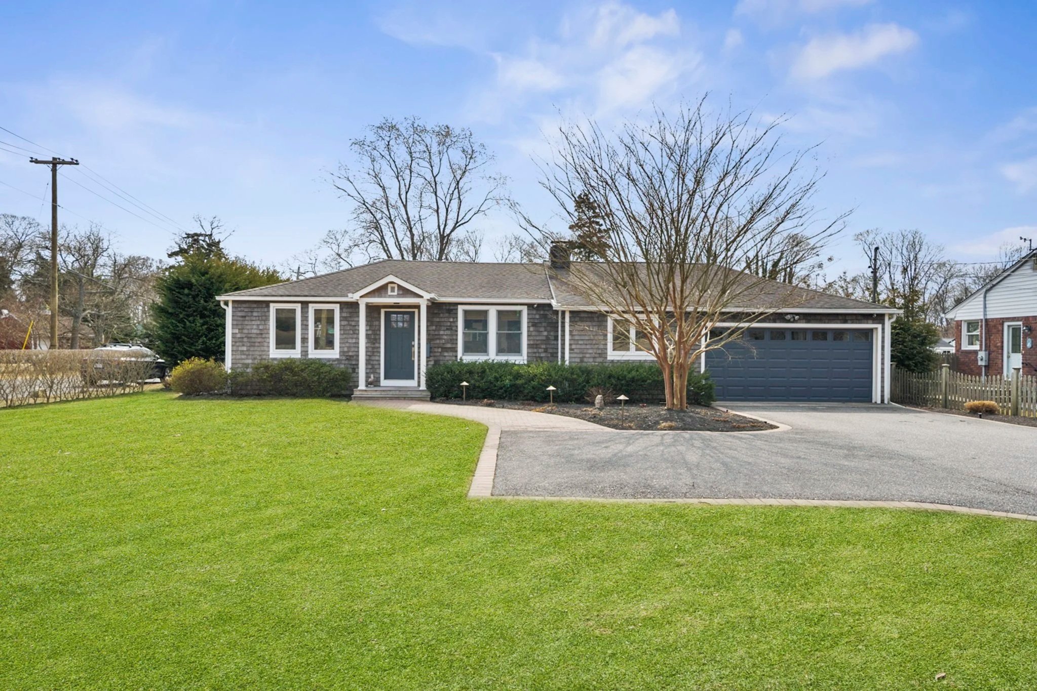 A single-story house with a gray shingle exterior and blue front door, surrounded by a well-maintained lawn and bushes. There are tall, leafless trees behind the house, a property fence on the right, and power lines to the left, under a partly cloudy blue sky.