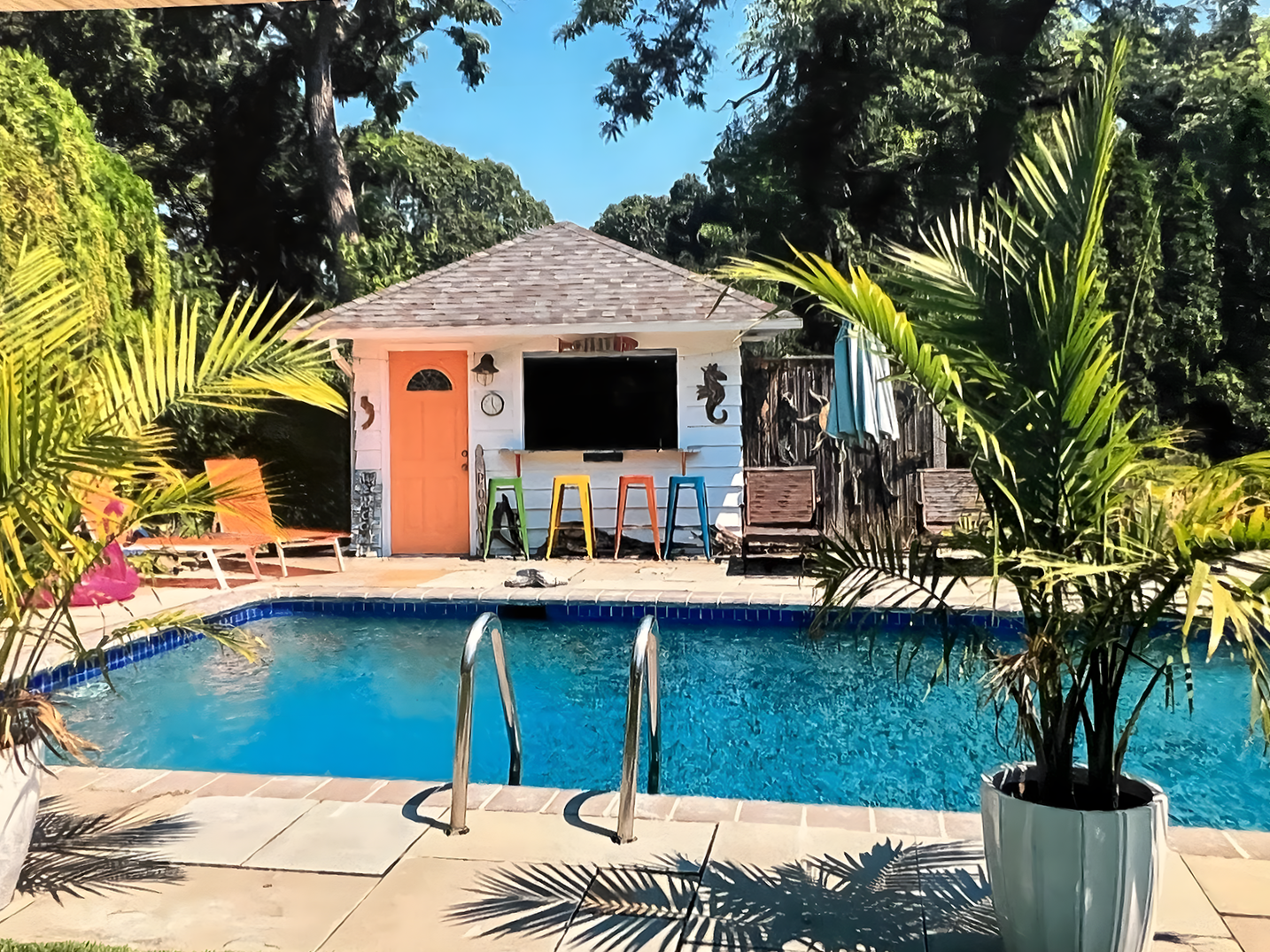 A backyard swimming pool with clear blue water, surrounded by a tiled deck, lush green plants, and trees. There is a small white shed with a gray shingled roof at the back, adorned with colorful stools, and outdoor furniture.