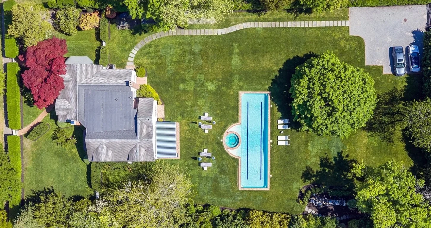 Aerial view of a backyard with a house, a swimming pool, a grassy lawn, trees, a stone pathway, and parked cars.