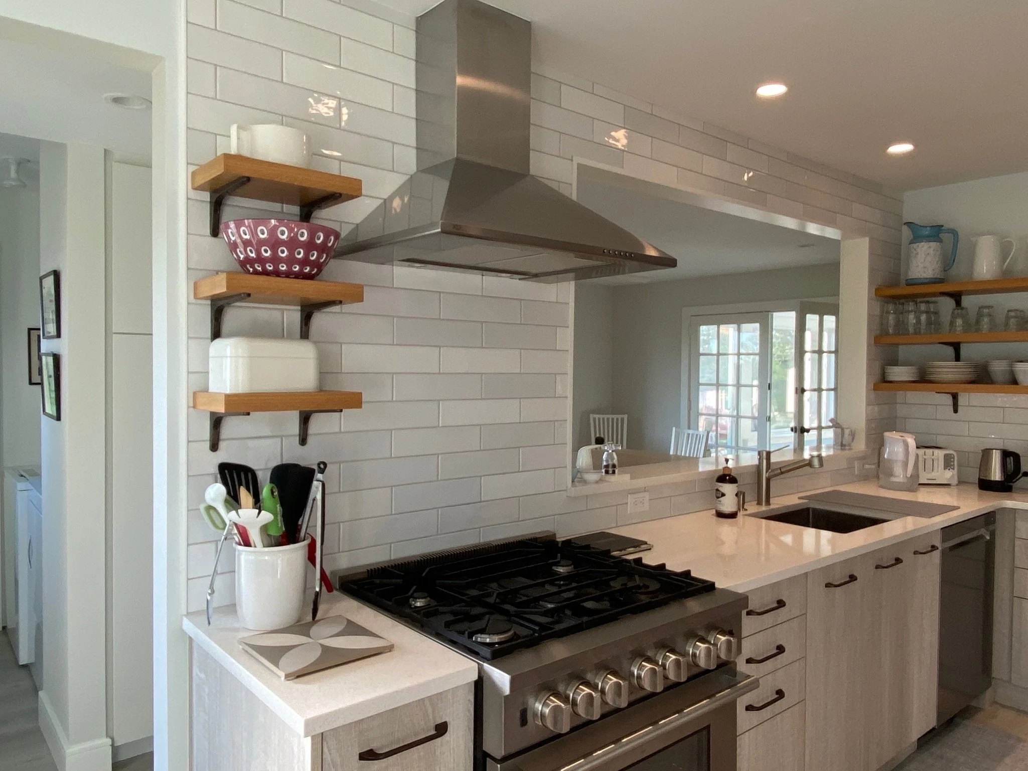 Modern kitchen with white brick backsplash, stainless steel range hood, black cooktop, open shelving with bowls and glassware, and a white marble countertop with kitchen utensils.