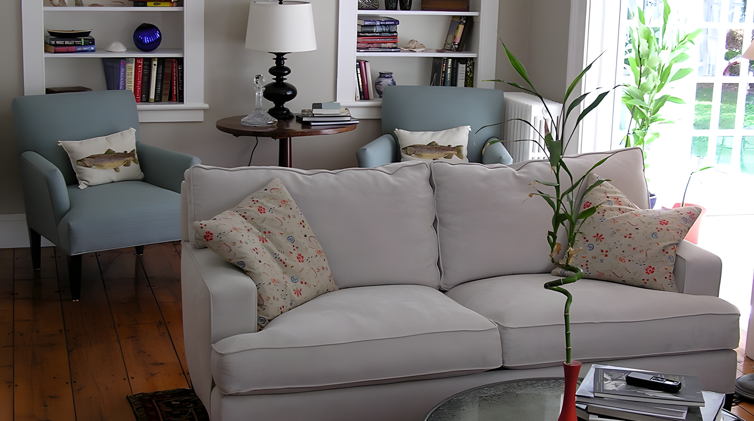 Living room with a white sofa, patterned pillows, a bamboo plant in a red pot, and a glass-top coffee table with books and a remote. Two light blue armchairs with fish pillows are positioned behind the sofa, with a round wooden side table and a black