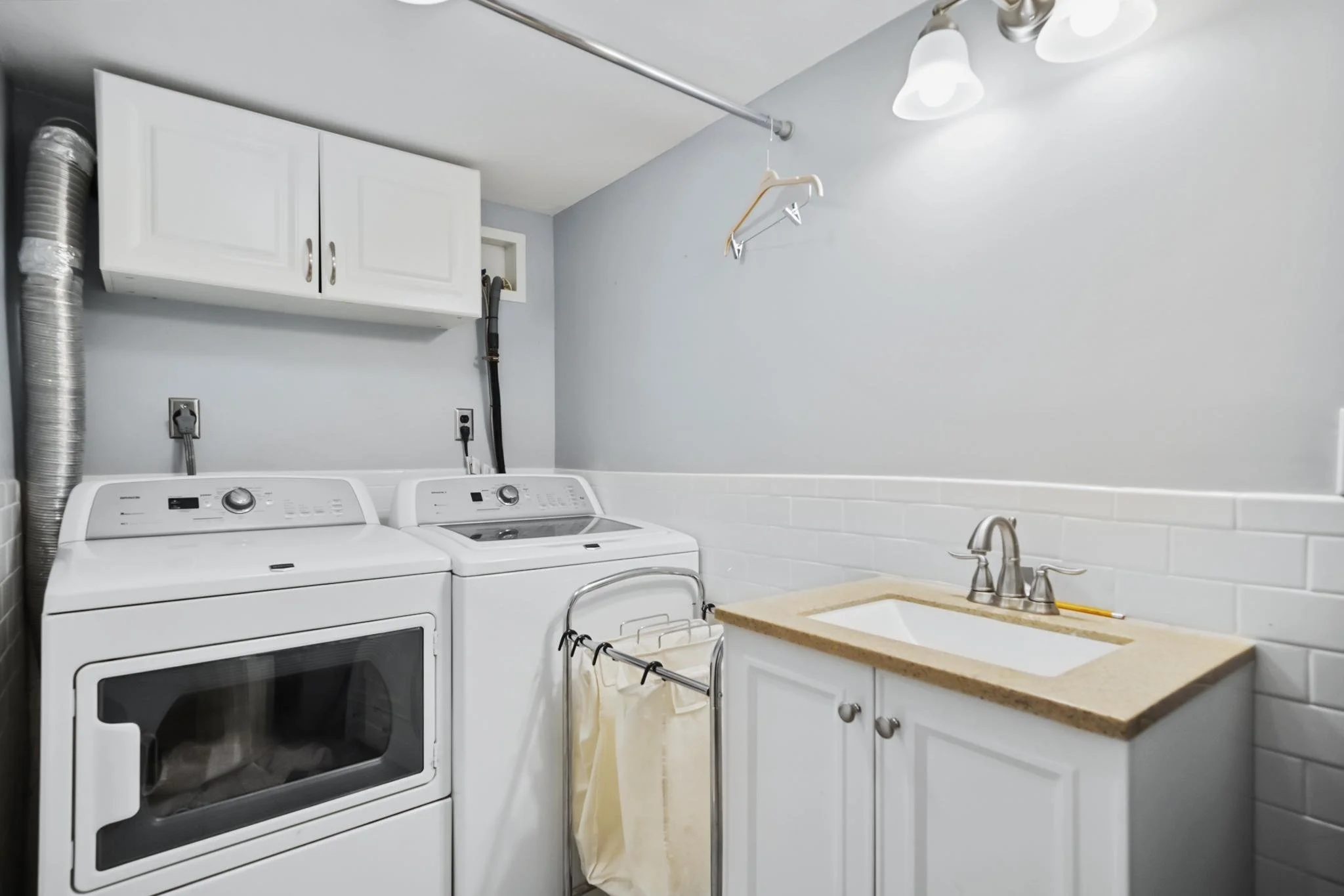 A laundry room with a white washing machine and dryer, white cabinets, a small sink, a beige countertop, and a clothes drying rod with an empty hanger.