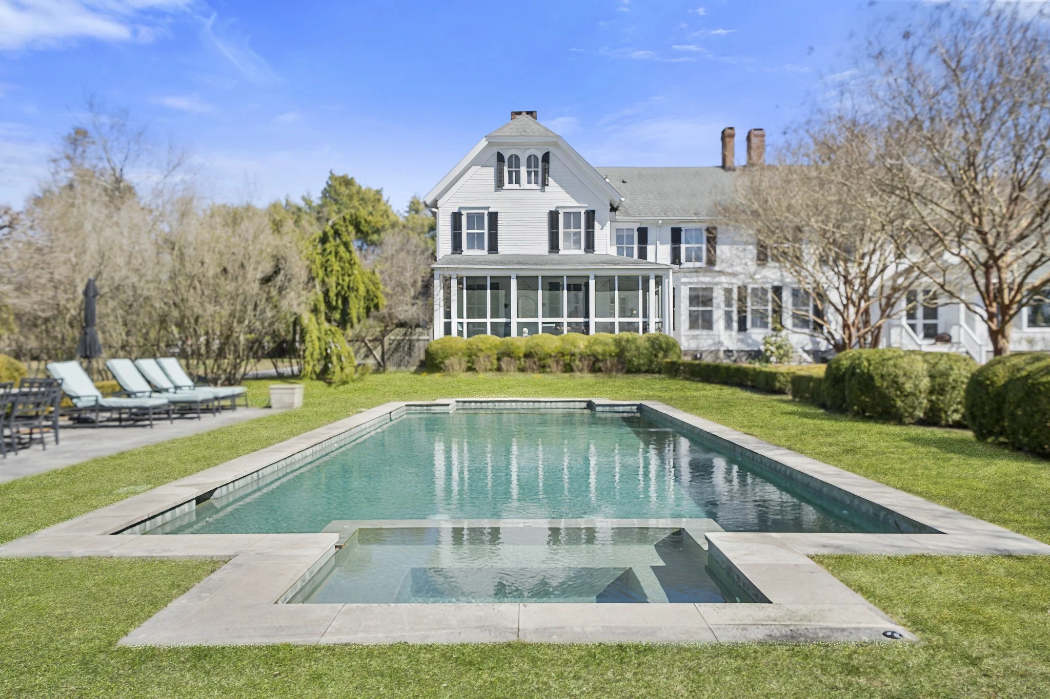 A backyard with a rectangular swimming pool, surrounded by a concrete deck and green grass. In the background, there is a large white house with black shutters and a screened porch, and trees with mostly bare branches.