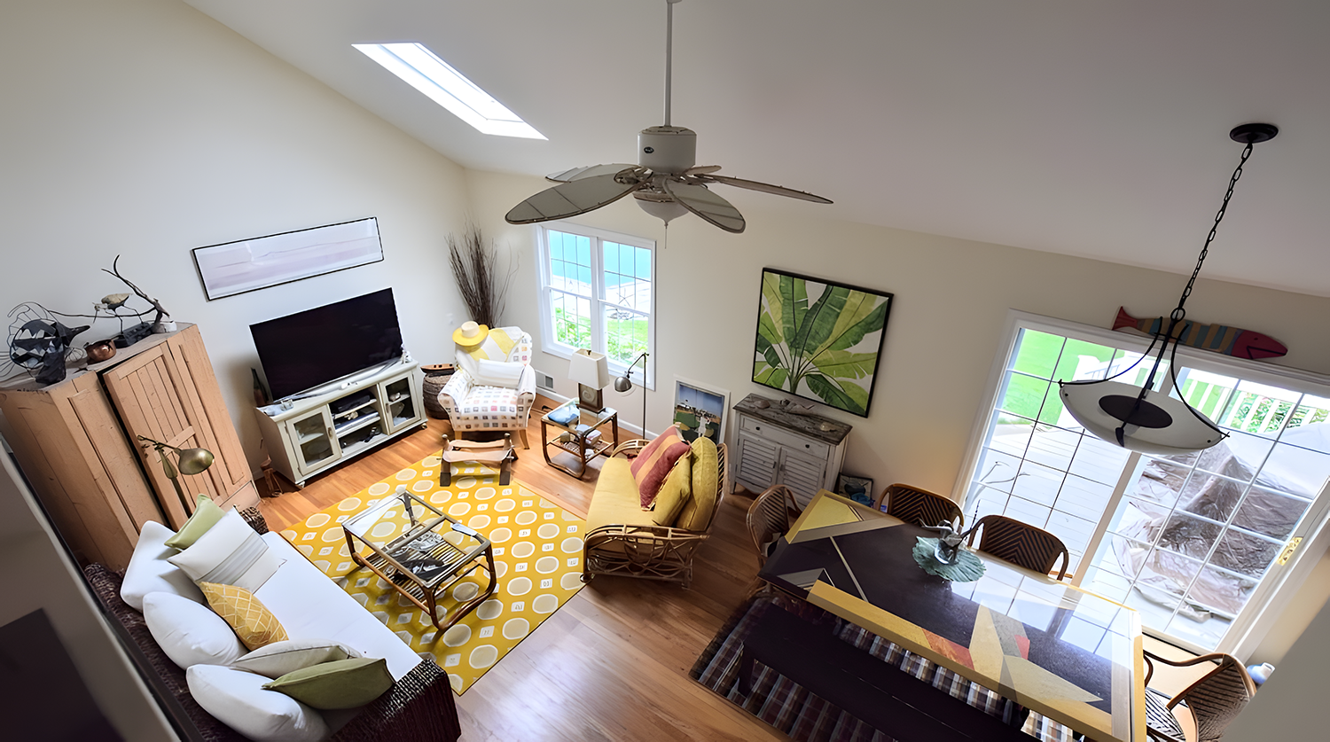 Living room with a high ceiling, ceiling fan, skylight, and large window, featuring a yellow and white patterned rug, white sofa, yellow armchair, a TV on a white stand, a wooden cabinet, and a dining table with chairs, decorated with plants and artw