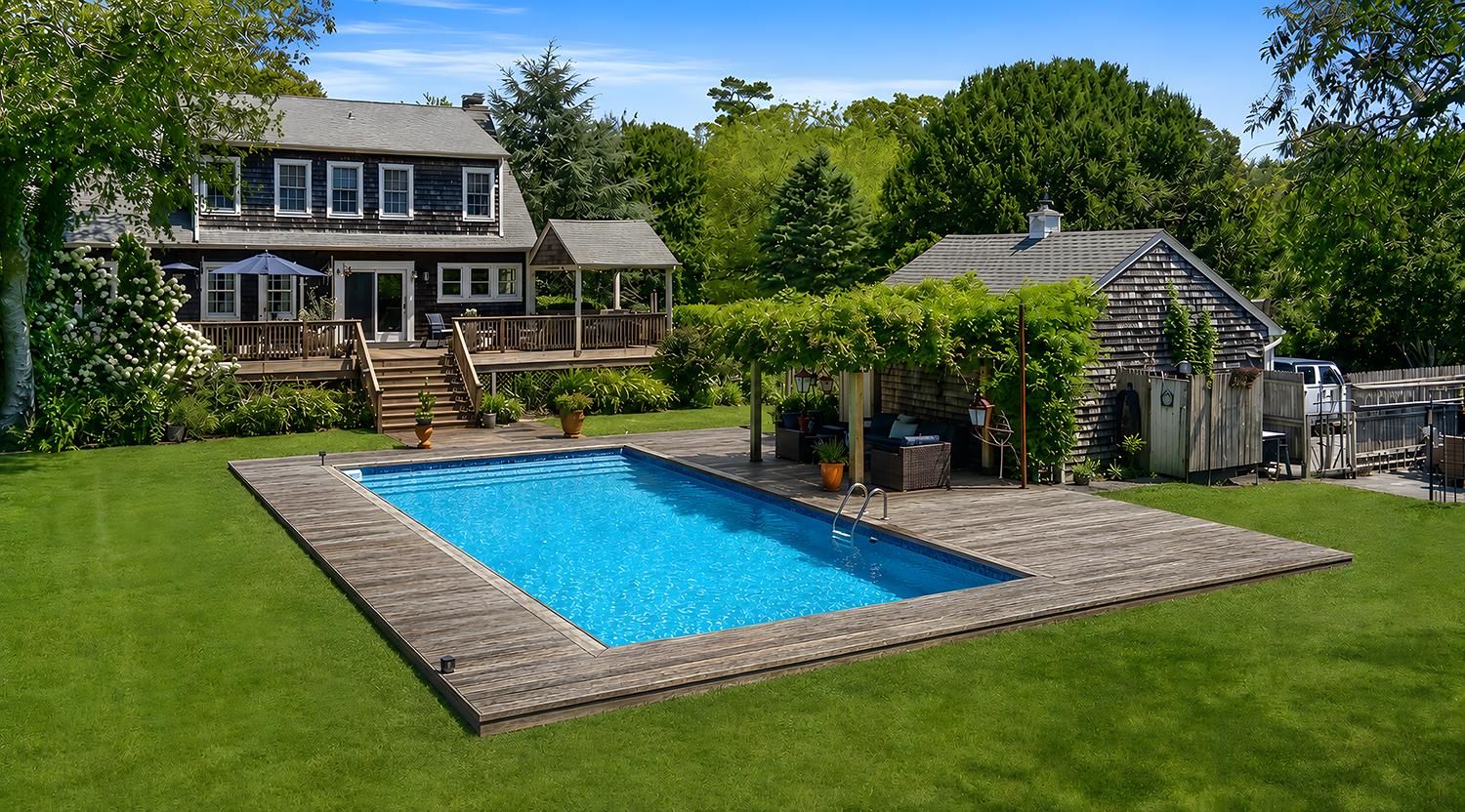 Backyard with a rectangular swimming pool surrounded by a wooden deck, green grass, a house with a deck, and a small shed covered with grapevines. There are trees and shrubs in the background.